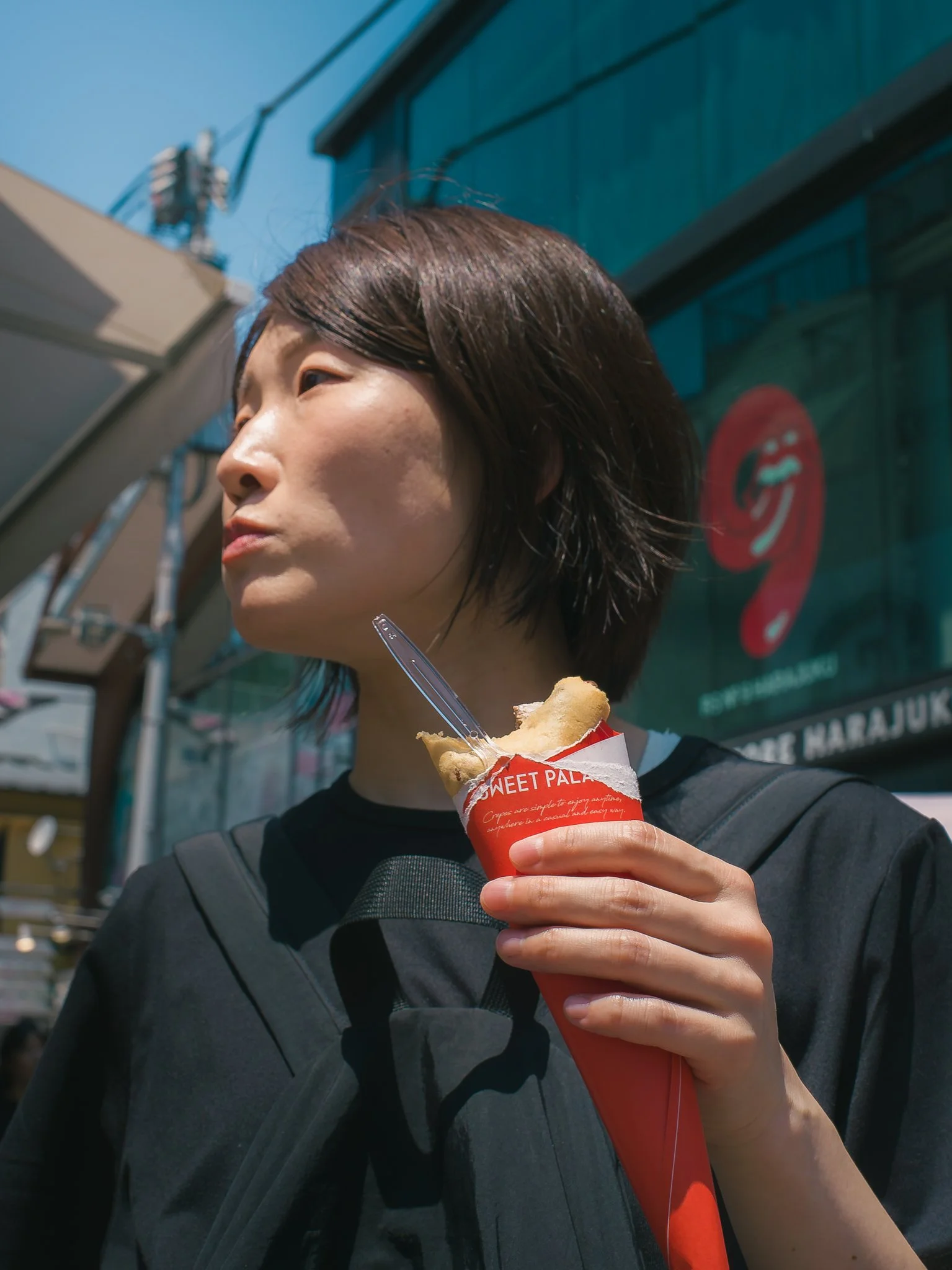 A woman with short dark hair holding a cone of ice cream in a red wrapper with the word "SWEET PALACE" written on it. She is wearing a black shirt and a backpack, standing outdoors on a sunny day, with buildings and a blue sky in the background.