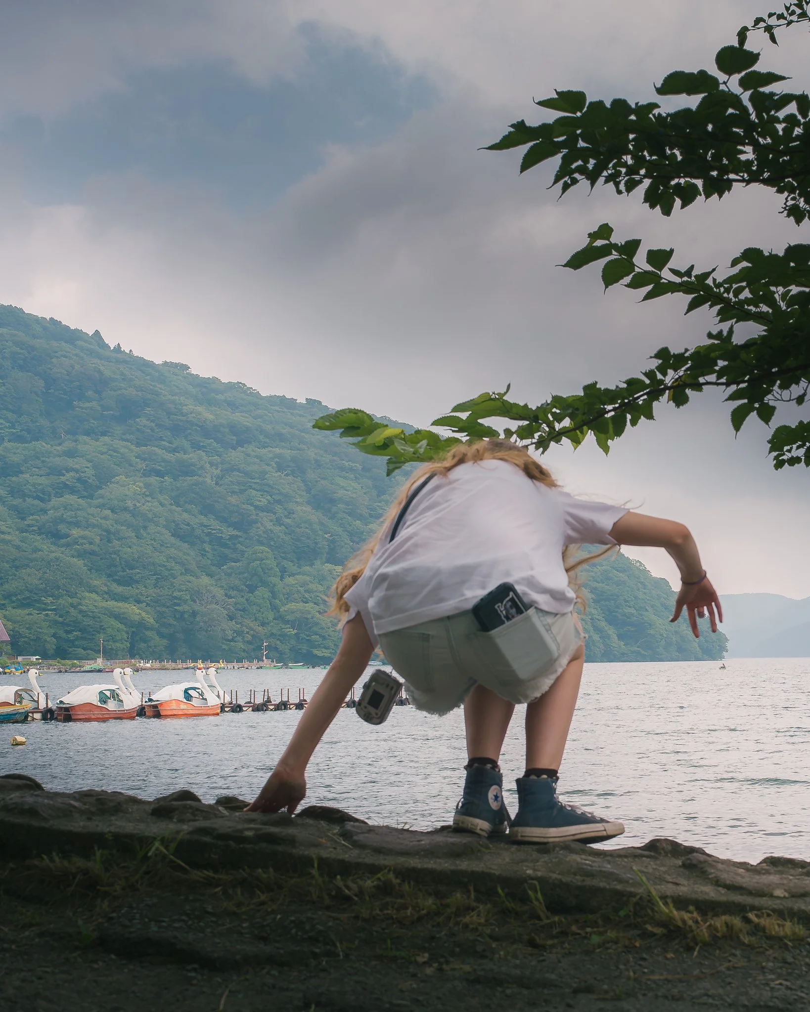 Person crouching by a lake shore with swan pedal boats in the water and green hills in the background on a cloudy day.