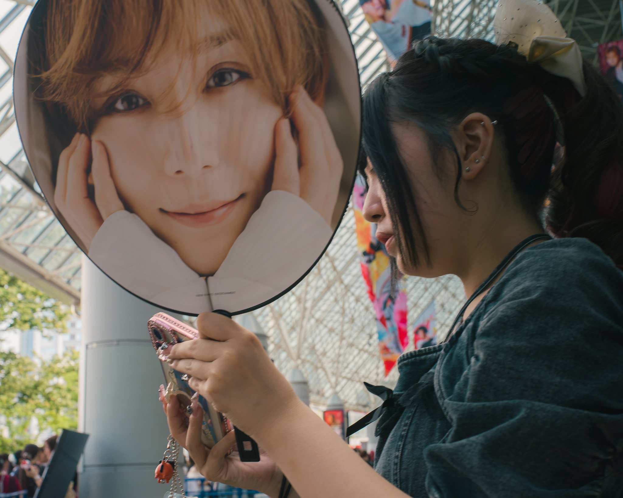 A woman outside at an event is holding a smartphone with both hands. She is wearing multiple earrings and has a flower in her hair. A large round fan with a printed photo of a young man with brown hair and striking eyes is in front of her face. In th