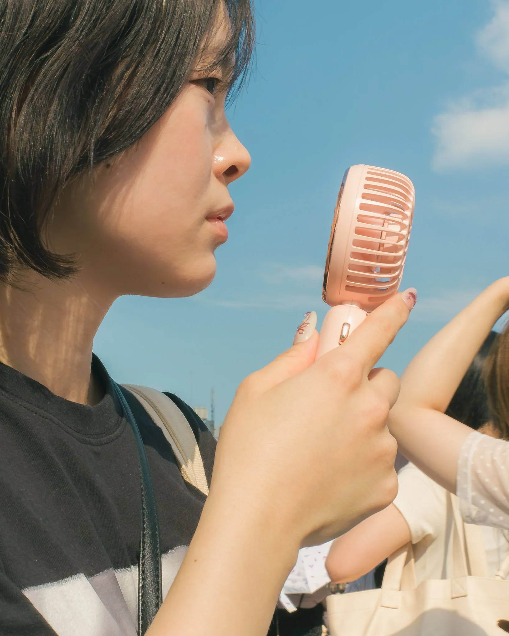 Close-up of a young woman with dark hair holding a pink handheld fan, sitting outdoors under a blue sky, with other people nearby.
