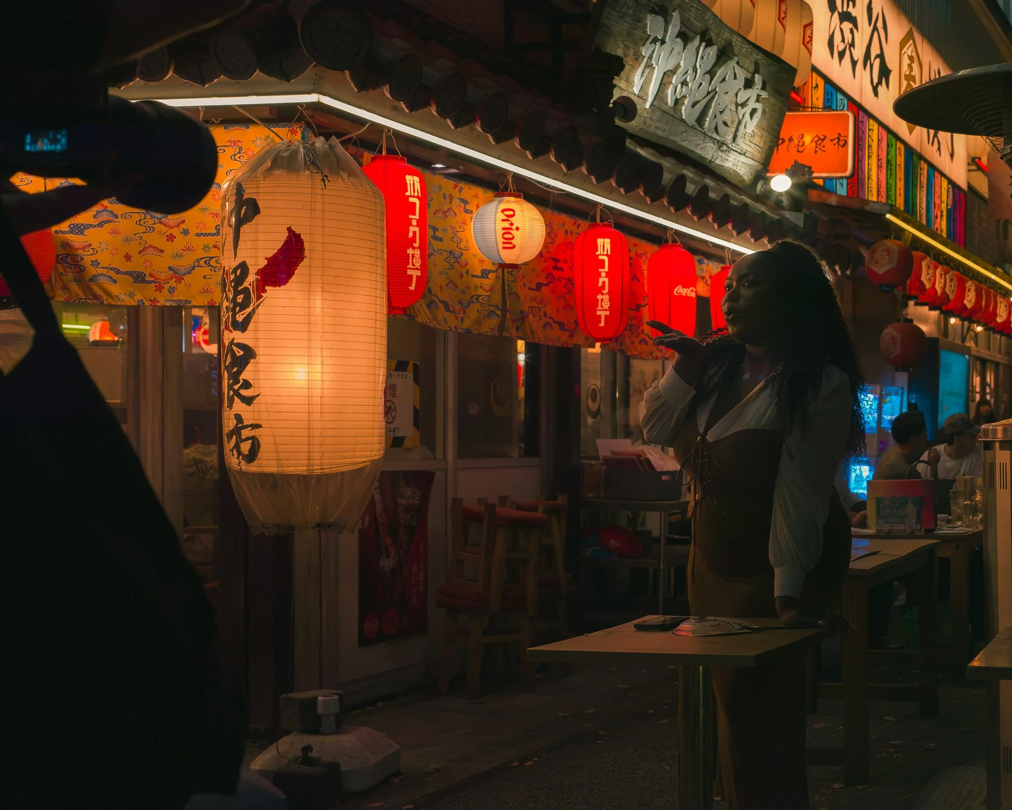 Night scene at a street market with Japanese lanterns and colorful signs, a woman with curly hair wearing a white top standing at a stall.