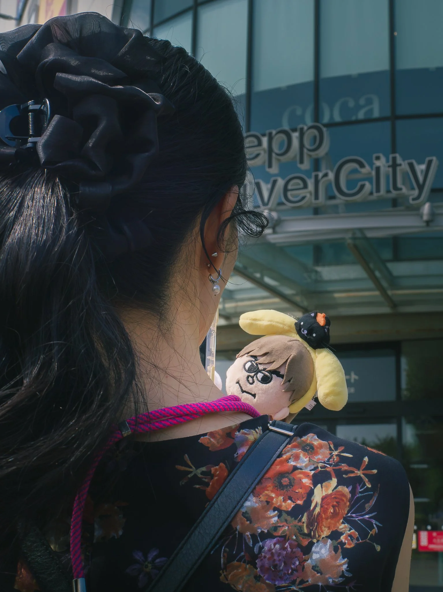 Close-up of a woman with long black hair tied with a large black scrunchie, wearing multiple earrings, holding a plush toy of a character with glasses and blonde hair, in front of a modern glass building with a sign reading 'Vancouver City'.