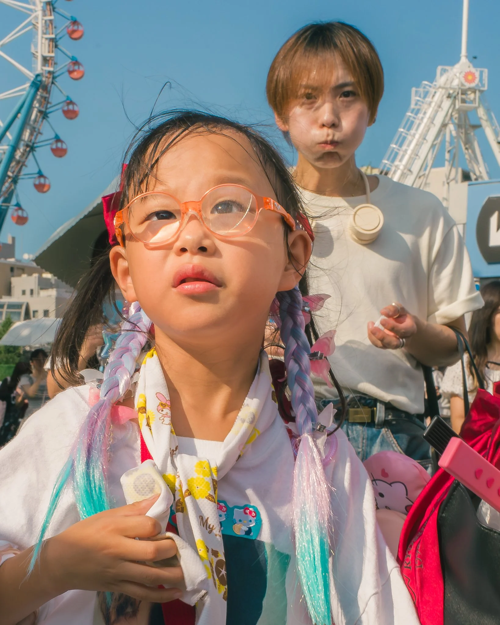 A young girl wearing pink glasses and colorful hair extensions holding an ice cream cone, with a woman and amusement park rides in the background on a sunny day.