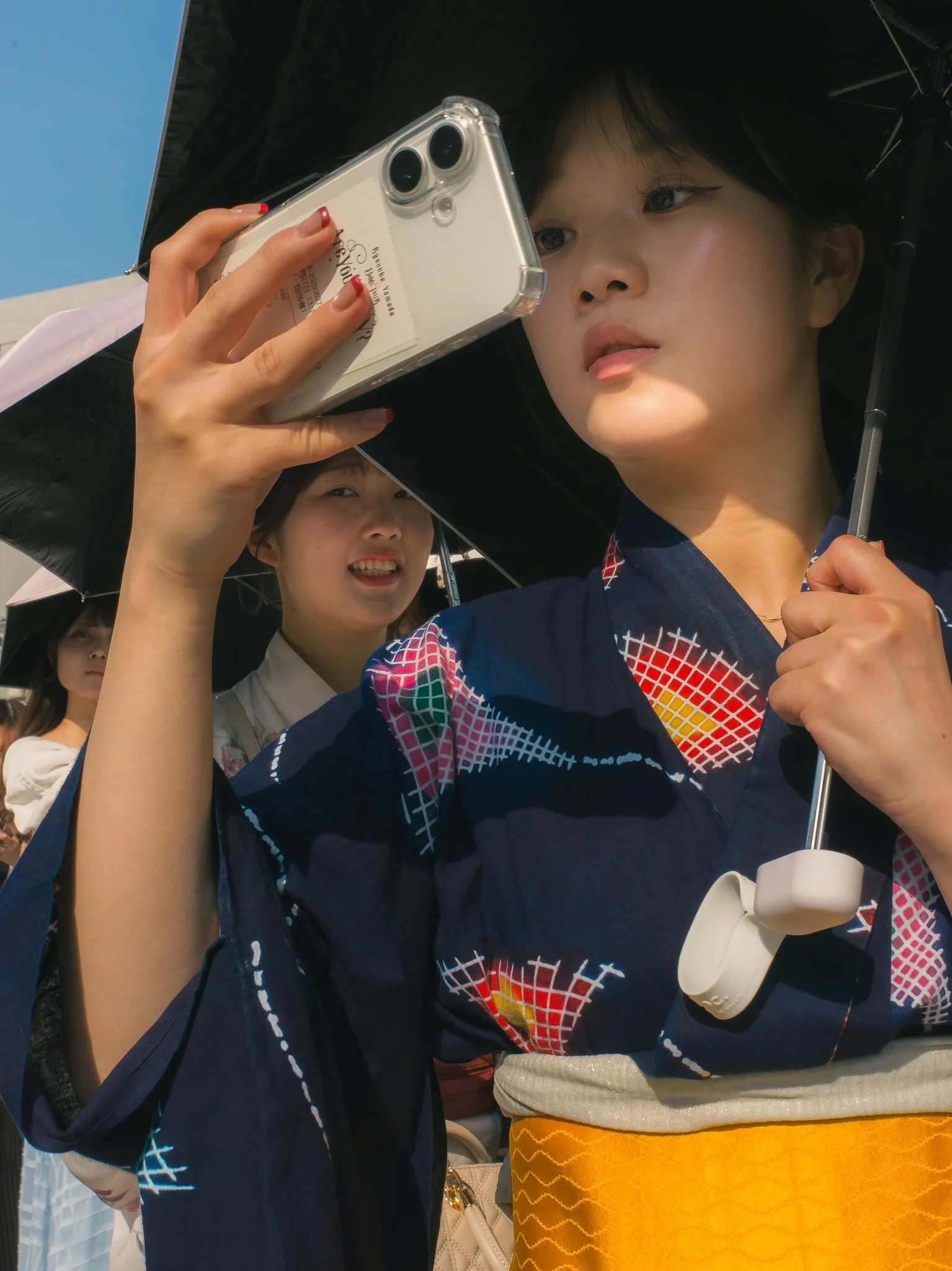Two women are outdoors, one in the foreground holding a smartphone close to her face, and another in the background smiling and looking. The woman in the foreground is wearing a colorful jacket and holding an umbrella. The woman in the background is 