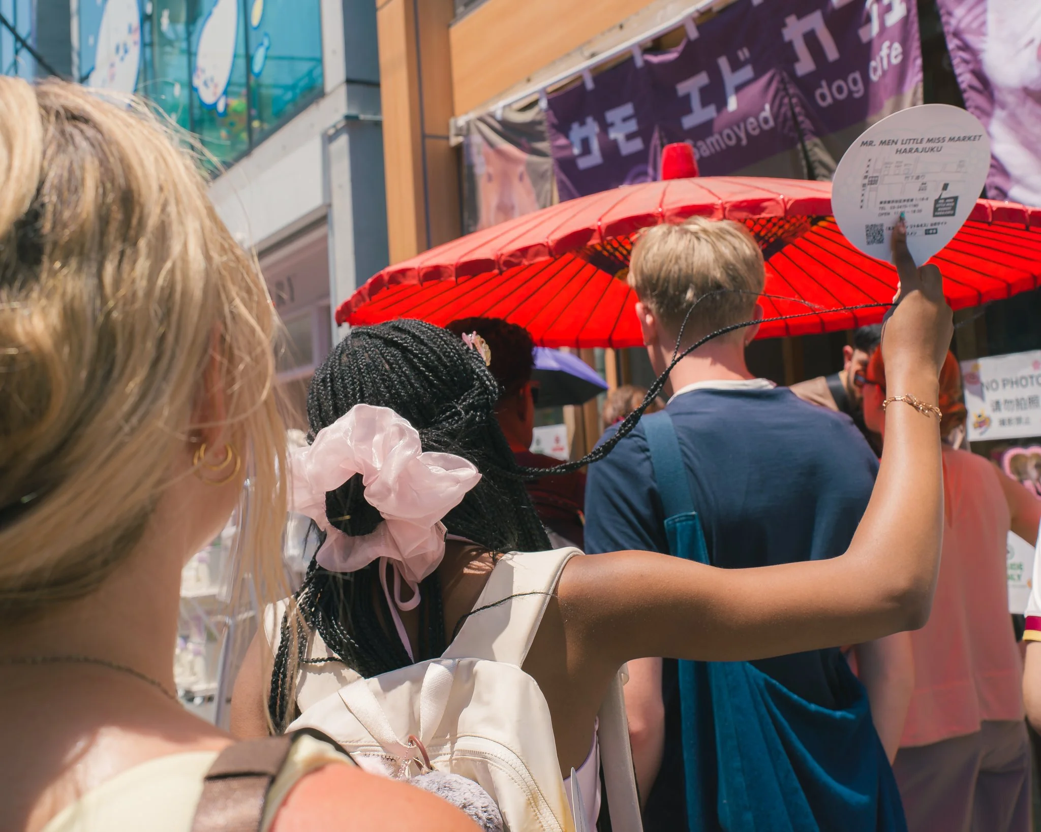 Crowd of people standing outdoors, some holding umbrellas and signs, at what appears to be a street event or parade, with buildings and banners in the background.