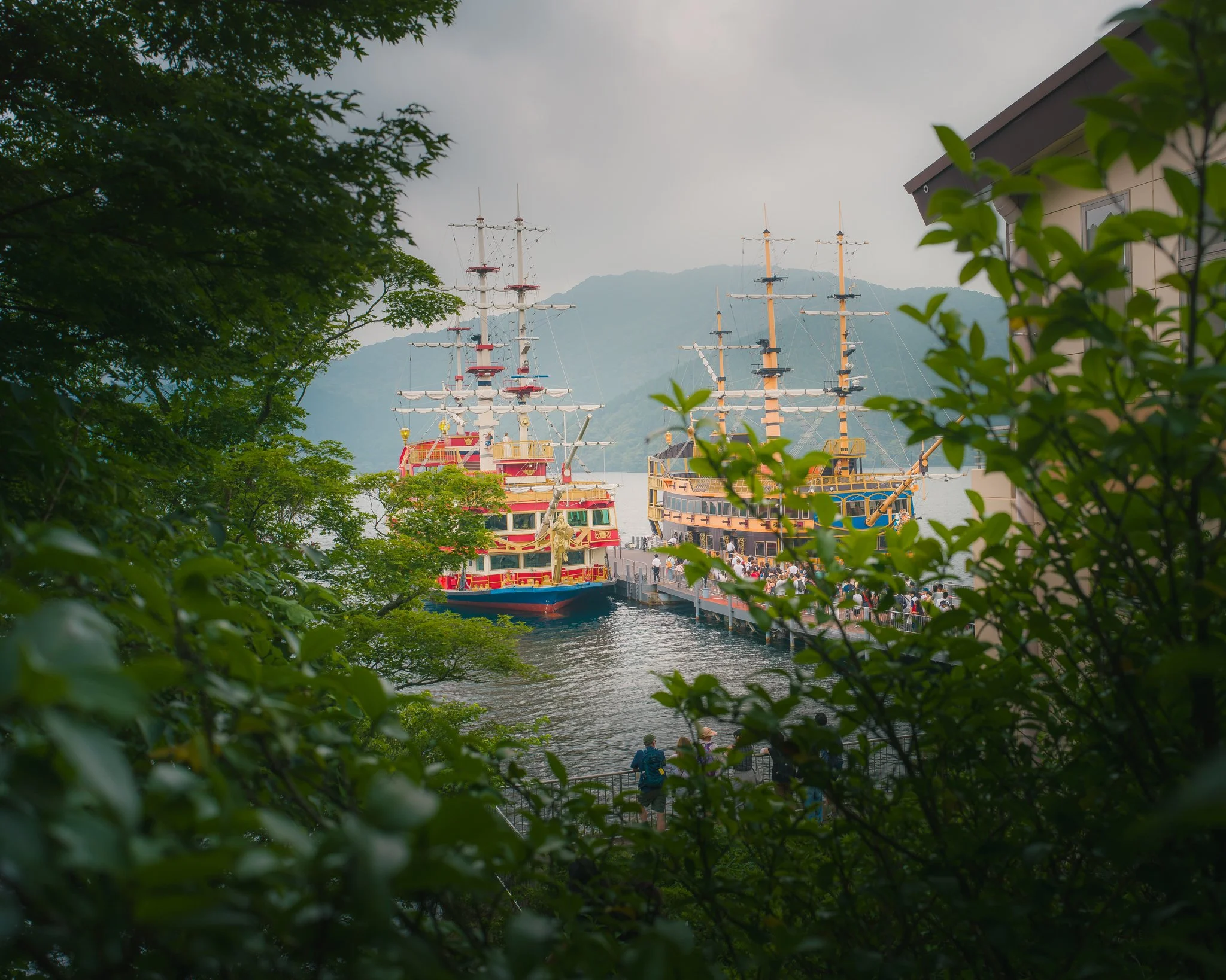 People walking near a docked pirate-themed ship with sails, trees, a building, and mountains in the background.