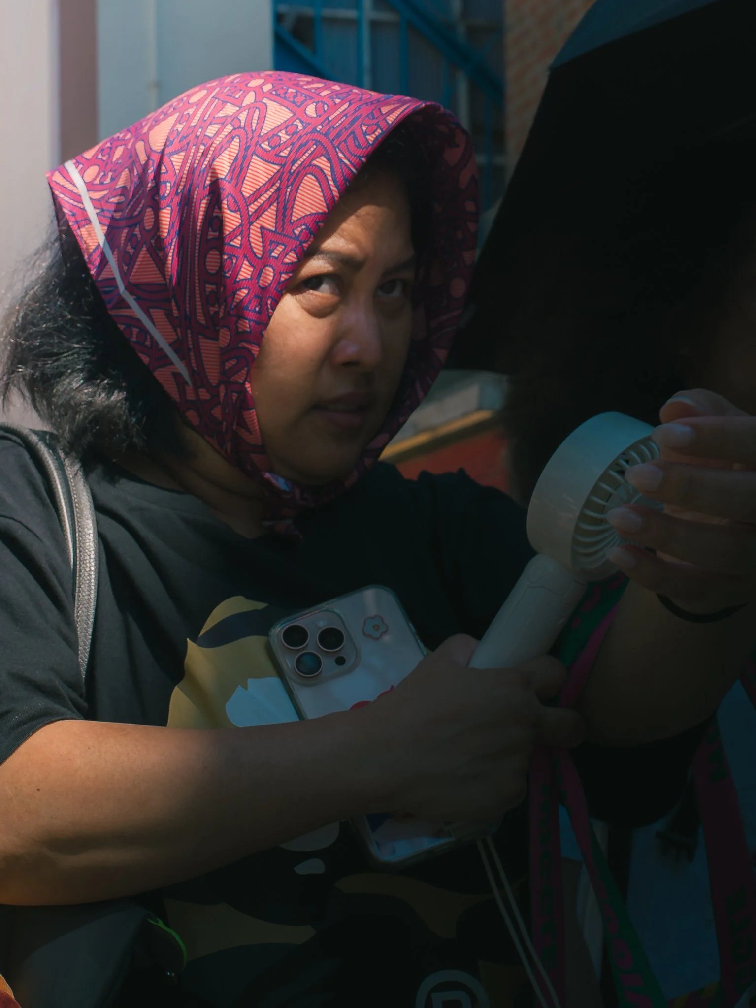 A woman with black hair wearing a colorful hooded jacket is holding a white handheld fan and a smartphone with a floral case. She appears to be outdoors.