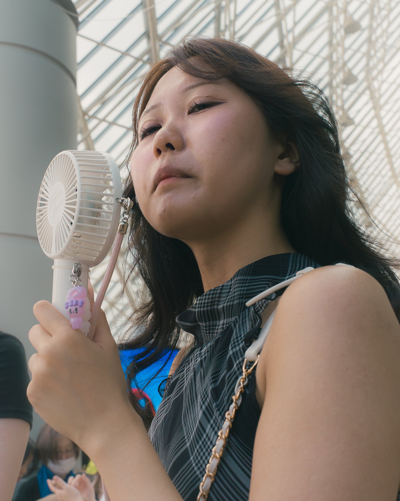 A woman with dark hair and makeup, wearing a black and white plaid shirt and gold chain purse, is using a handheld fan with a pink beaded keychain. She appears to be indoors under a glass ceiling.