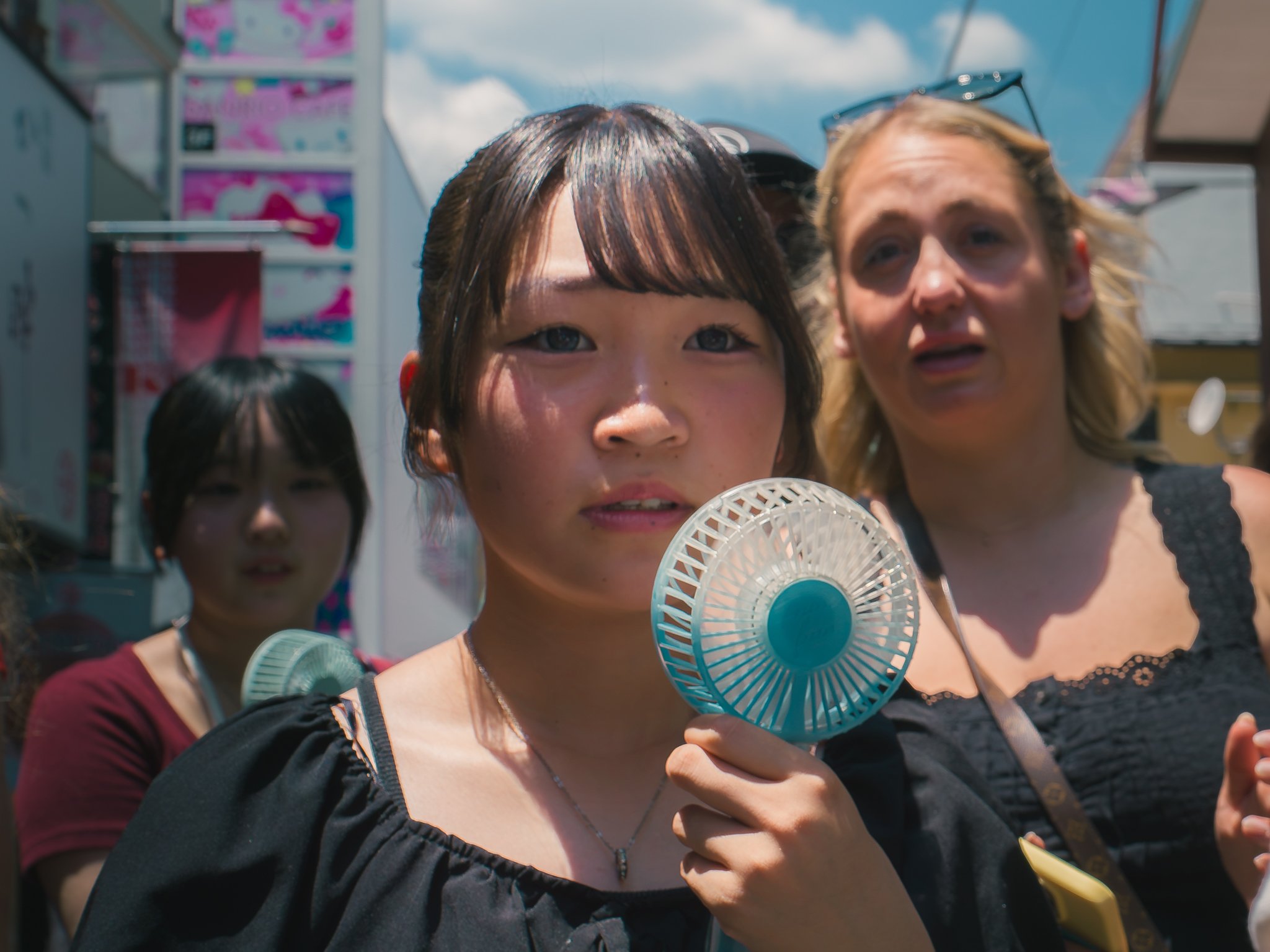 A group of people outside in a sunny day, with a woman in the foreground holding a small blue fan, and other women behind her, with some wearing sunglasses and carrying bags.