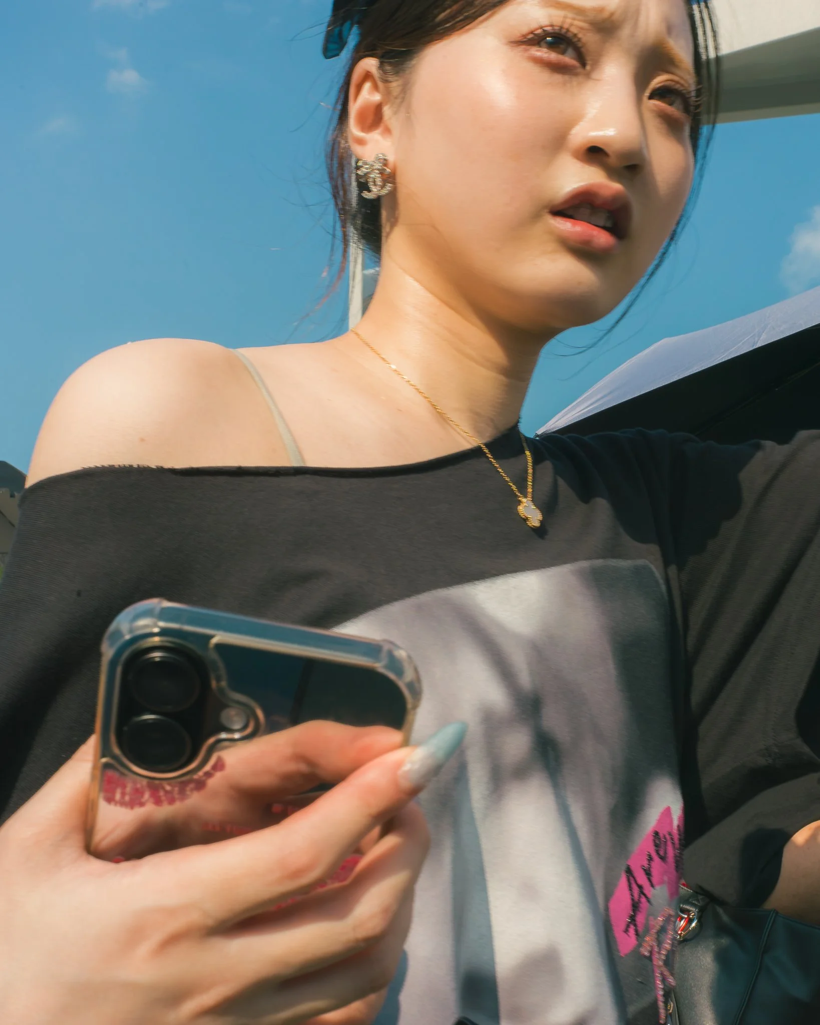 A woman with earrings and a necklace taking a selfie with a smartphone, with a blue sky in the background.