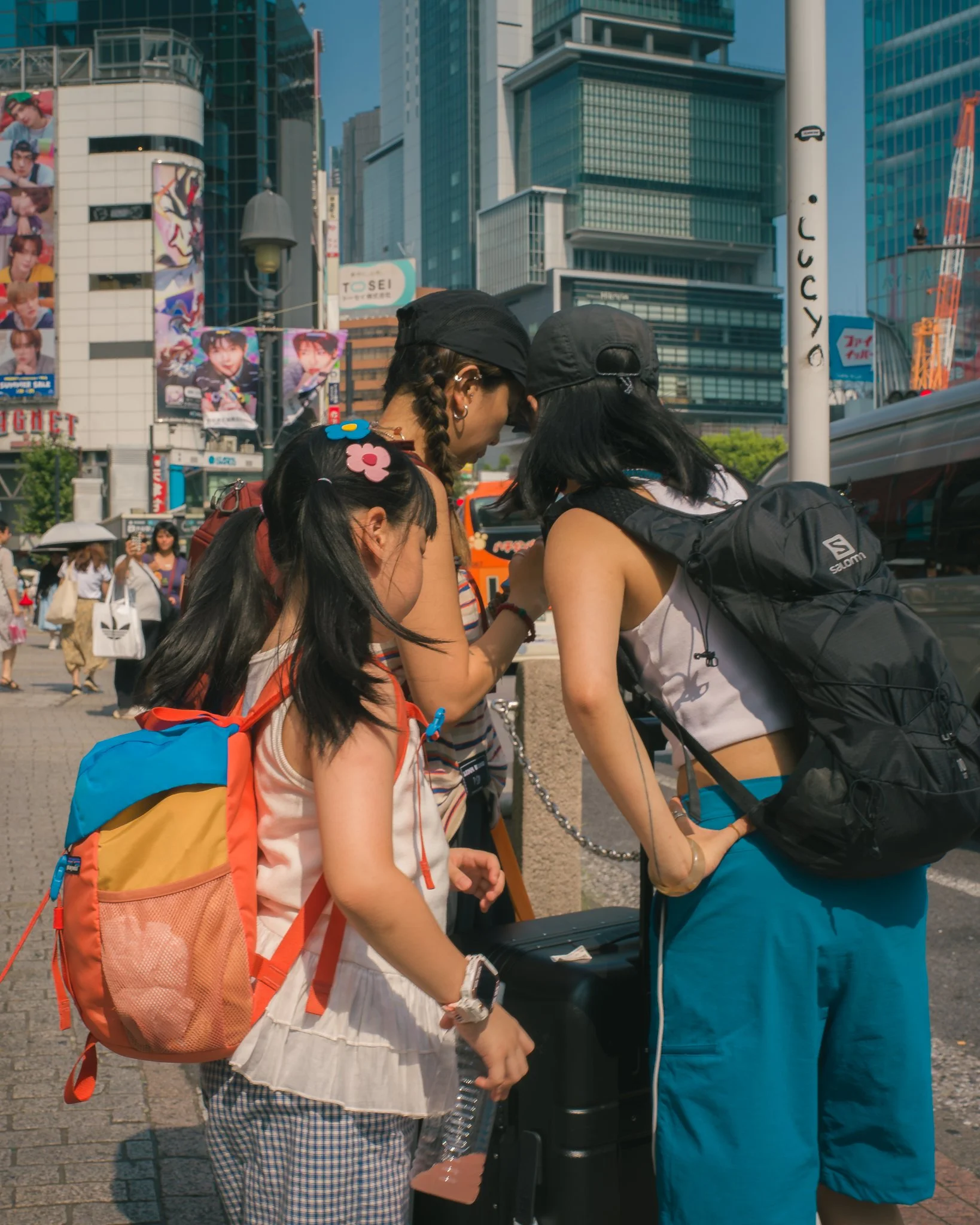 Three young women standing together on a busy city street, wearing casual summer clothes, backpacks, and hats, engaged in a conversation near a black trash bin, with tall modern buildings and colorful posters in the background.