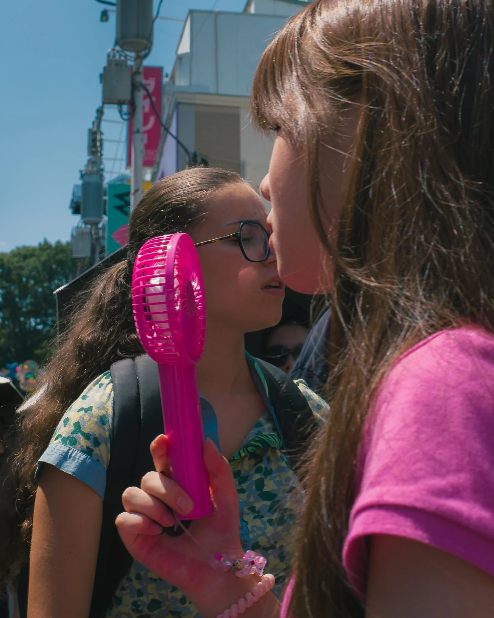 A woman and young girl are sharing a close moment, with their faces touching. The girl is wearing glasses and holding a pink fan. They are outdoors, with a building and power lines in the background.