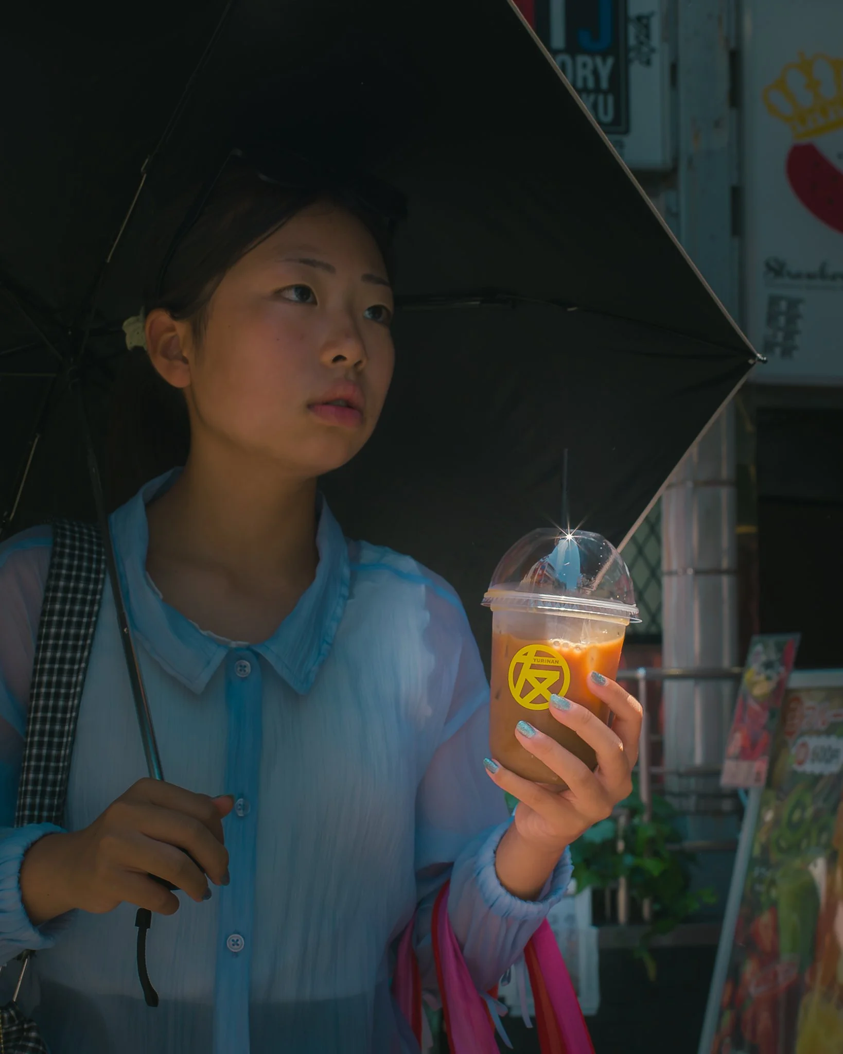 A woman holding a plastic cup with an iced coffee beverage and a black umbrella, standing near a street stall or shop on a sunny day.