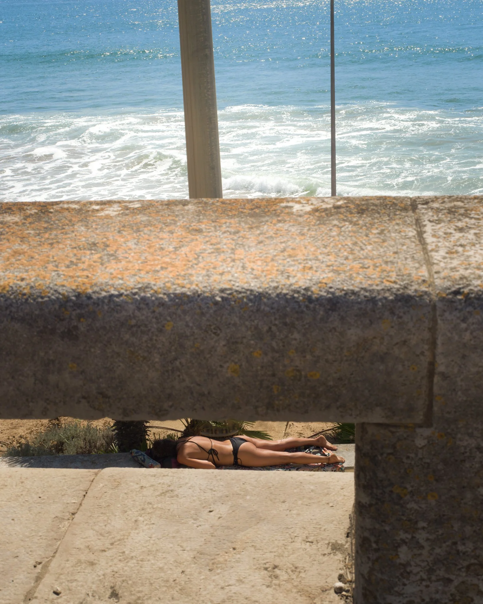 A woman in a black swimsuit lying on a towel at the beach, viewed through gaps in a stone or concrete wall.