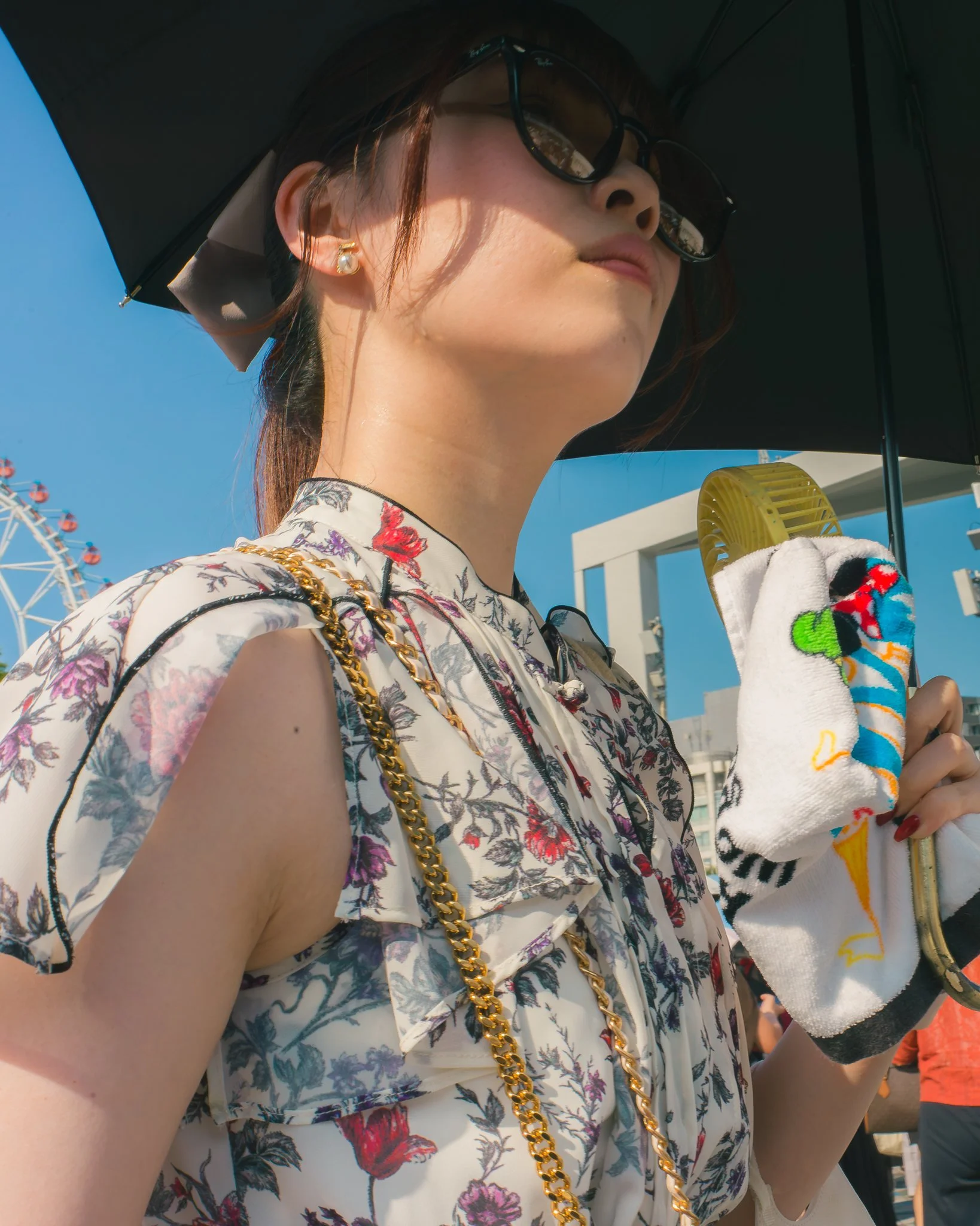 Close-up of a woman wearing sunglasses, pearl earrings, a floral dress, a black umbrella, and holding a colorful towel at an amusement park with a Ferris wheel in the background.