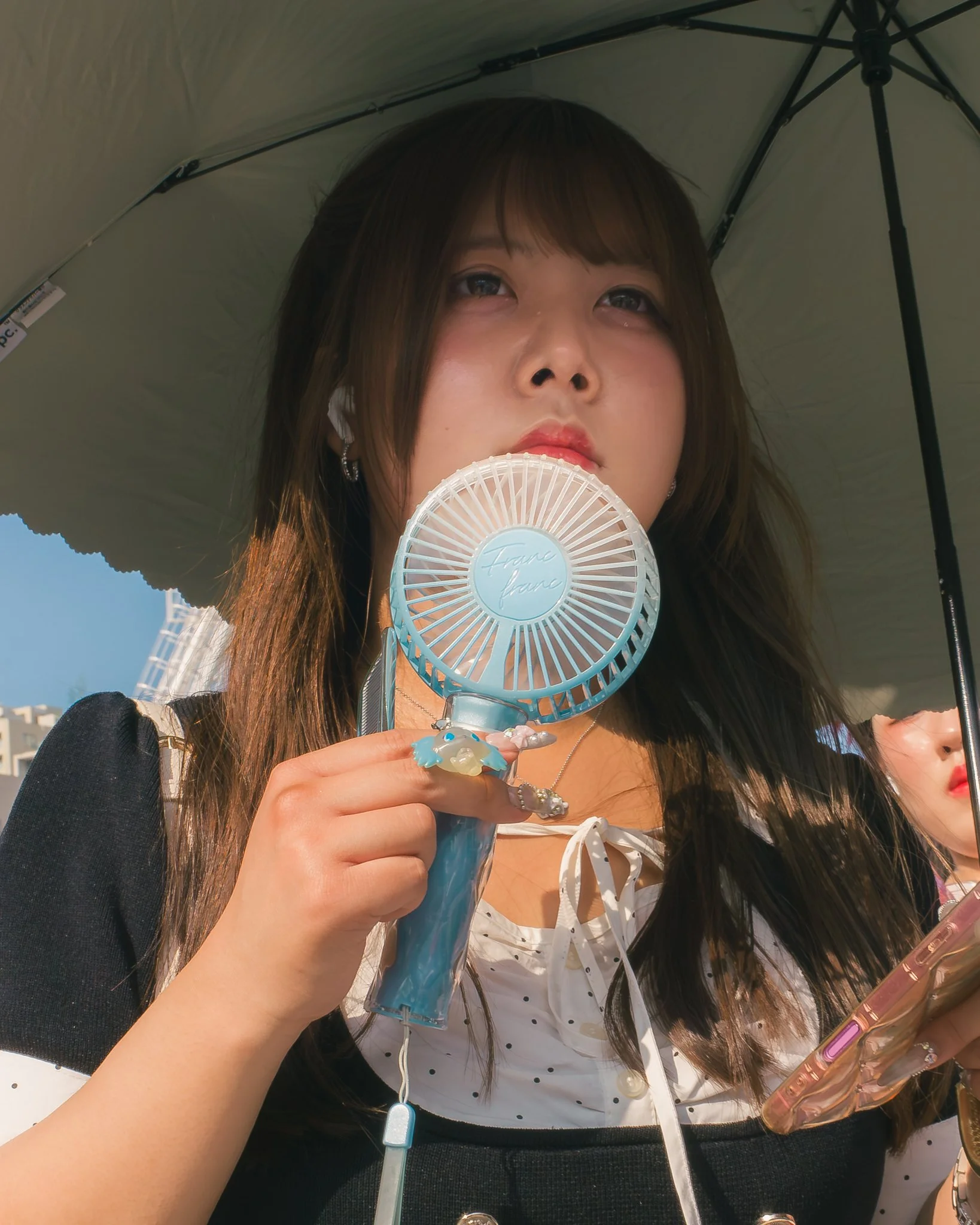 Close-up of a woman holding a blue handheld fan, with long dark hair, wearing earrings and a polka-dot top, shielding herself with a large umbrella against sunny weather.