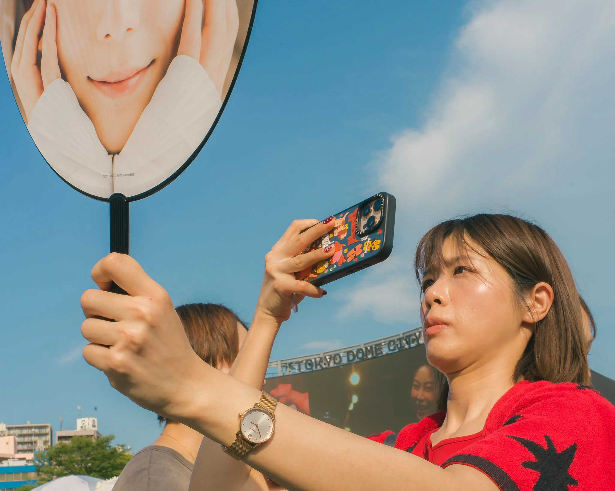 Two women are taking a selfie while holding a large mirror with a woman's face on it. One woman is wearing a red and black striped shirt and a gold wristwatch, and is smiling. The other woman is partially visible, holding a colorful smartphone with a