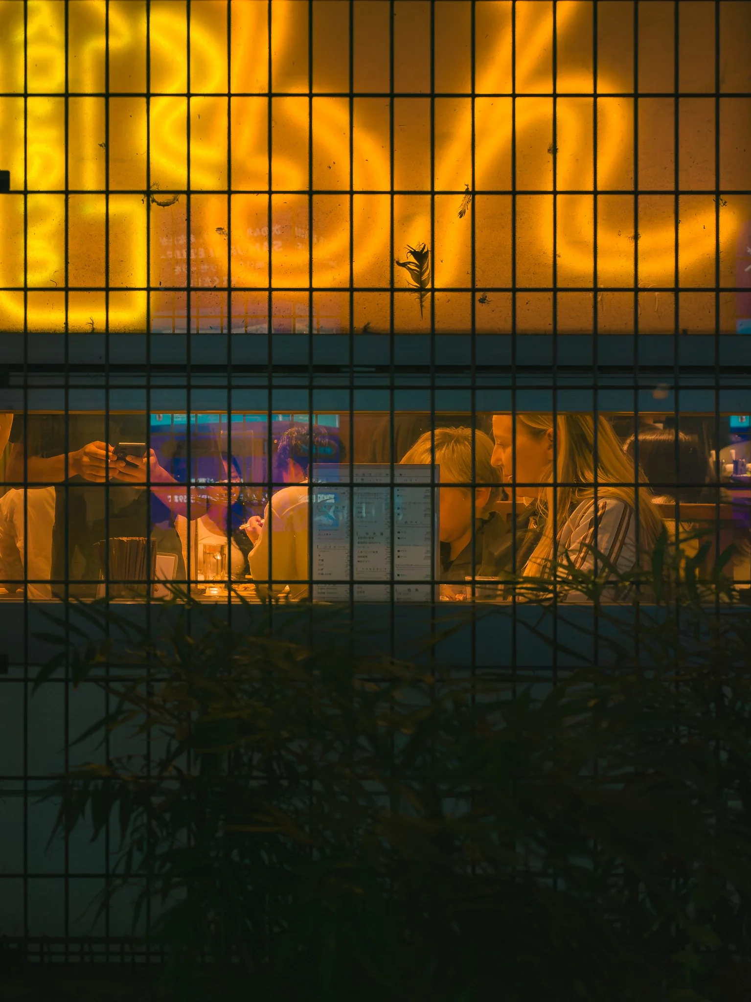 View through a wire fence of people at a dining table inside a restaurant with a yellow illuminated sign on the wall behind them.