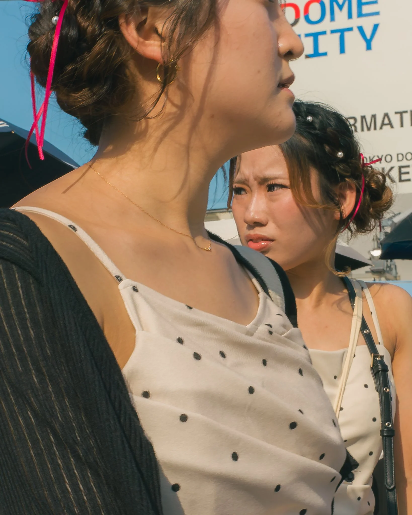 Close-up of two young women with curly hair, one in the foreground wearing a black top with sheer striped sleeves and a white tank top with black polka dots, and the other in the background with a white top, both with earrings, against a backdrop wit