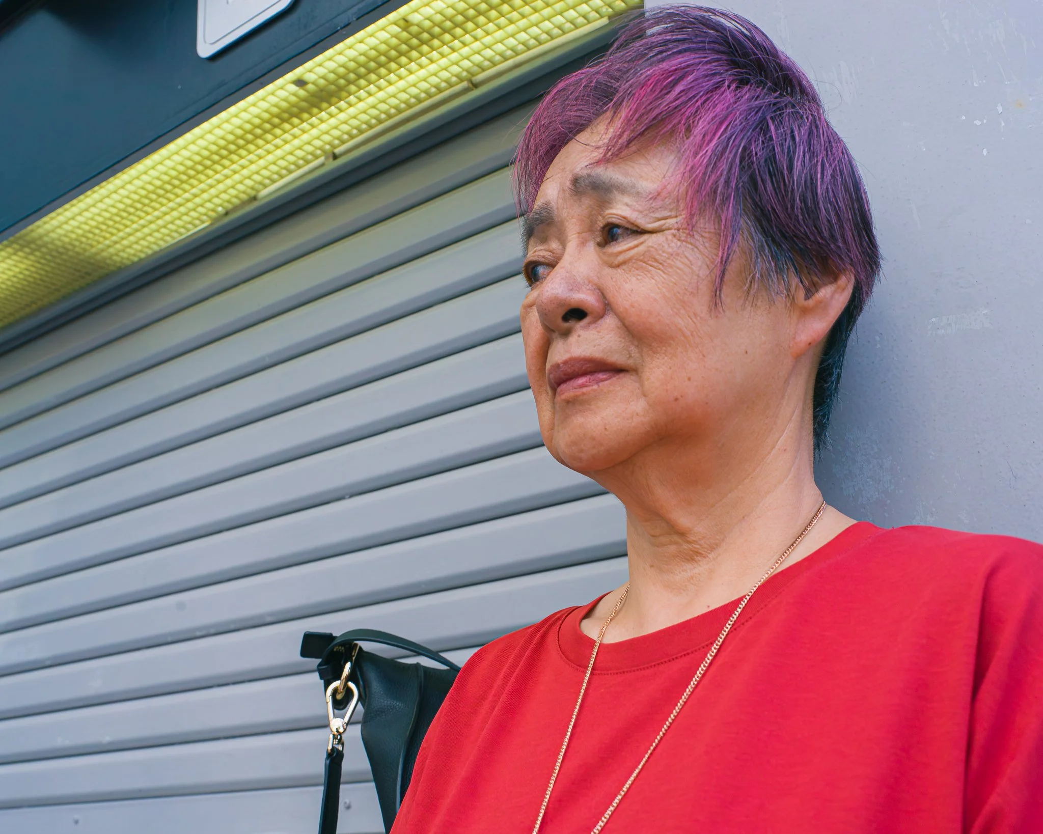 A woman with short purple hair wearing a red shirt and gold necklace standing against a gray metal wall with yellow lighting.