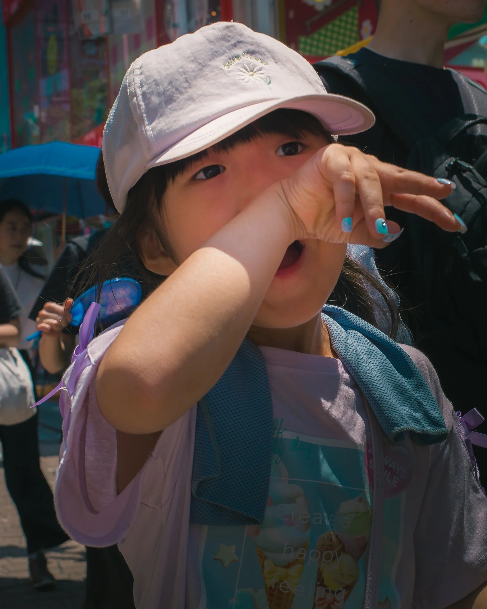 A young girl wearing a white cap and backpack, holding her hand to her nose while standing outdoors at a busy area with colorful signs and people in the background.