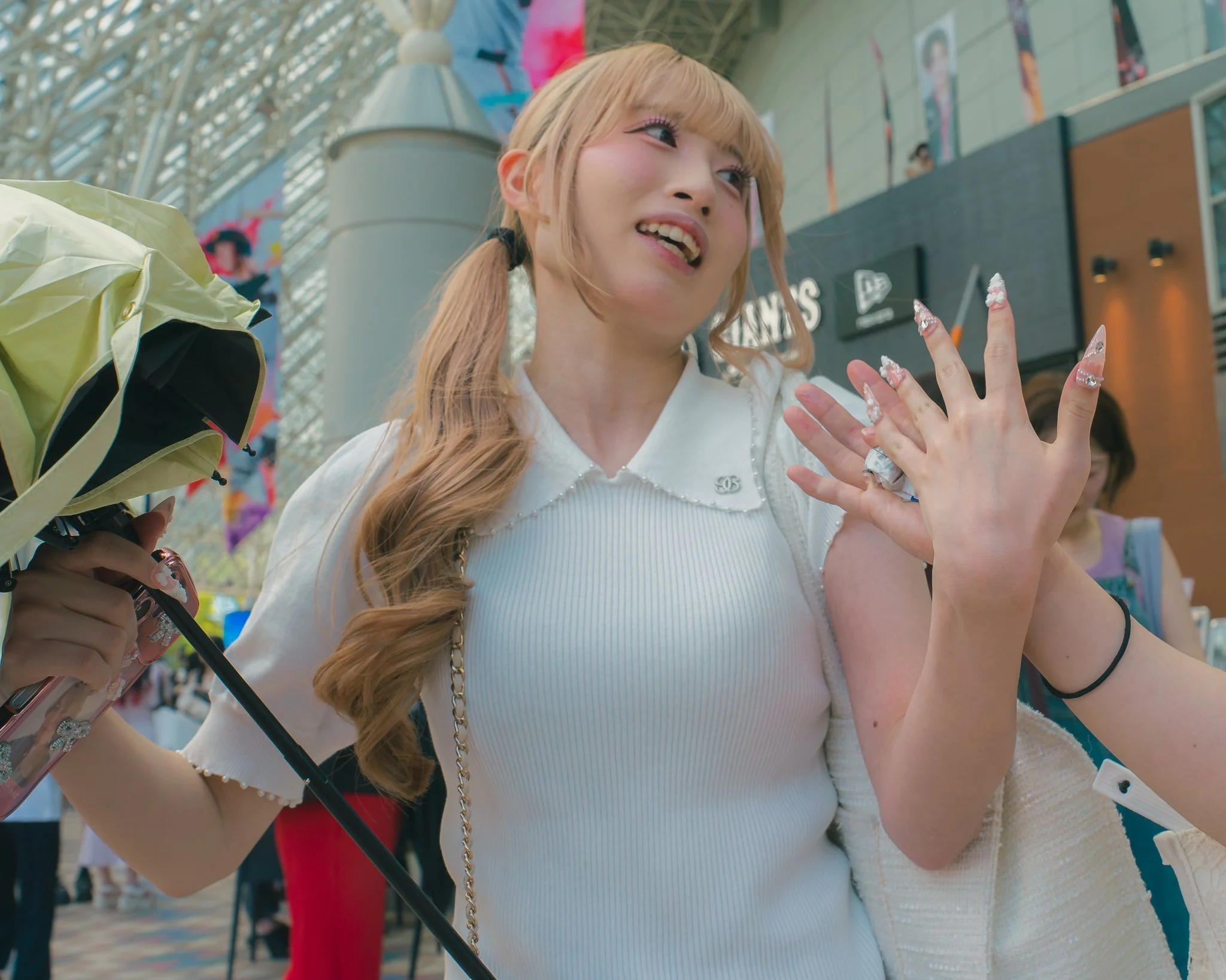 Close-up of a young woman with long blonde hair in pigtails, smiling and showing off her decorated fingernails at what appears to be an indoor amusement park or entertainment venue.
