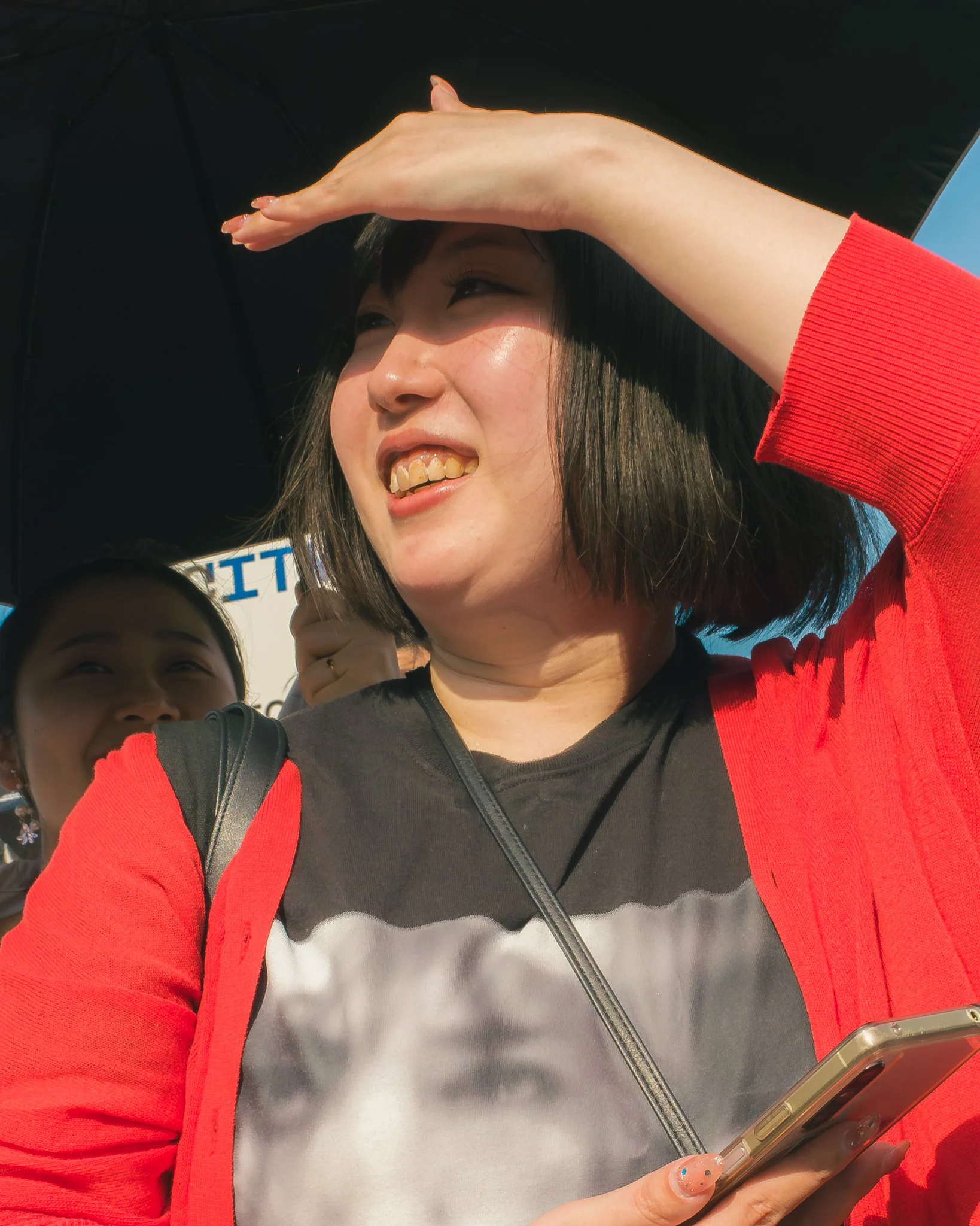A woman with short dark hair and light skin, wearing a red jacket over a black and white shirt, holding a smartphone with a transparent case, and shielding her eyes from sunlight with her hand while smiling.