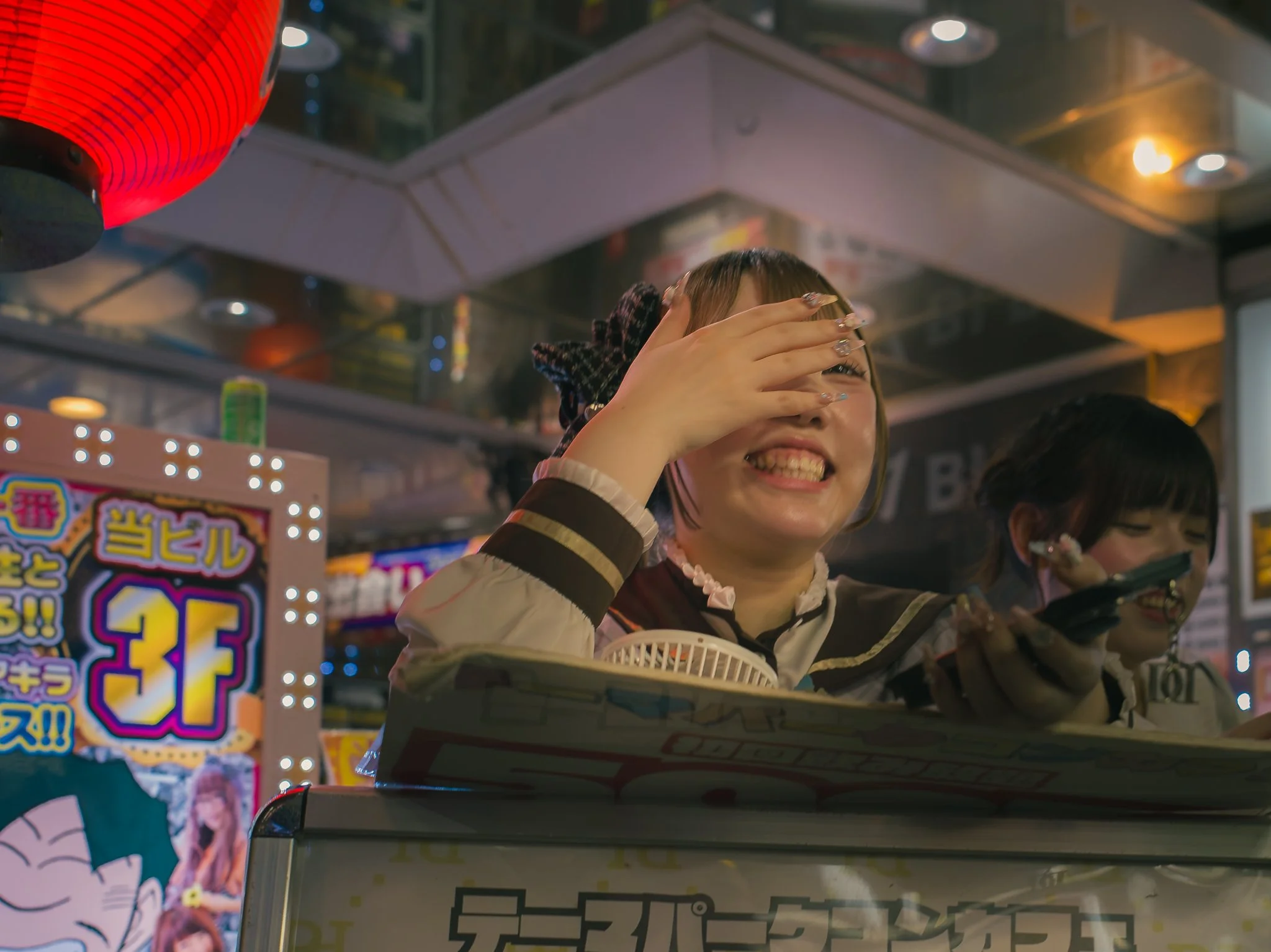 A smiling woman with dark hair and bangs covering her eyes, wearing a black and white striped top with a necklace, at a lively carnival or arcade, with colorful signs and a red lantern overhead.