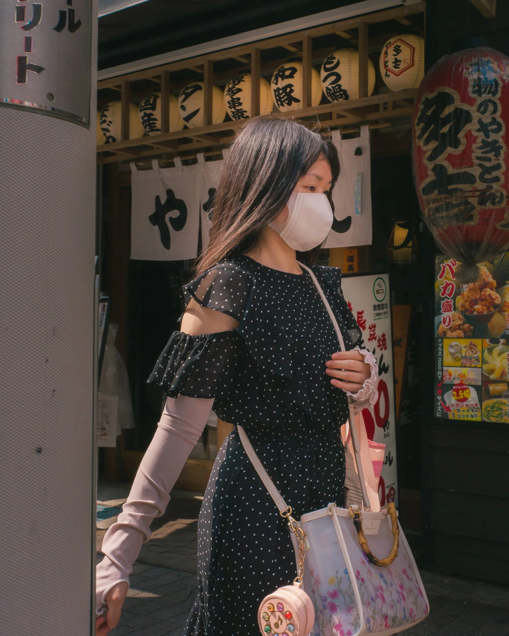 A young woman with long dark hair, wearing a white face mask, a black dress with white polka dots, and beige sleeves, is standing on a city street. She is carrying multiple handbags, including a light pink bag with a colorful circular embellishment, 