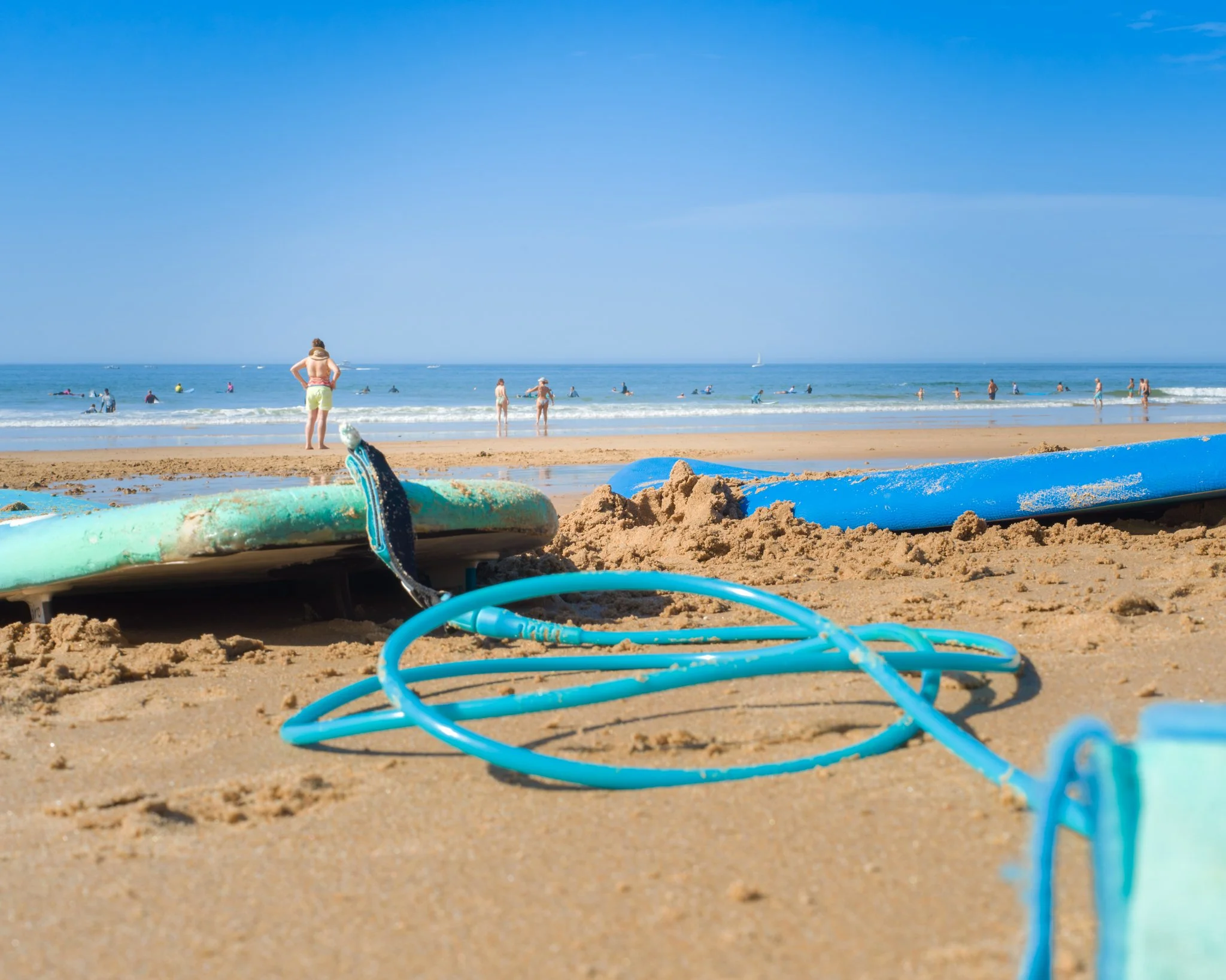 Sandcastle and blue hose on sandy beach with people swimming and walking in the ocean under clear blue sky.
