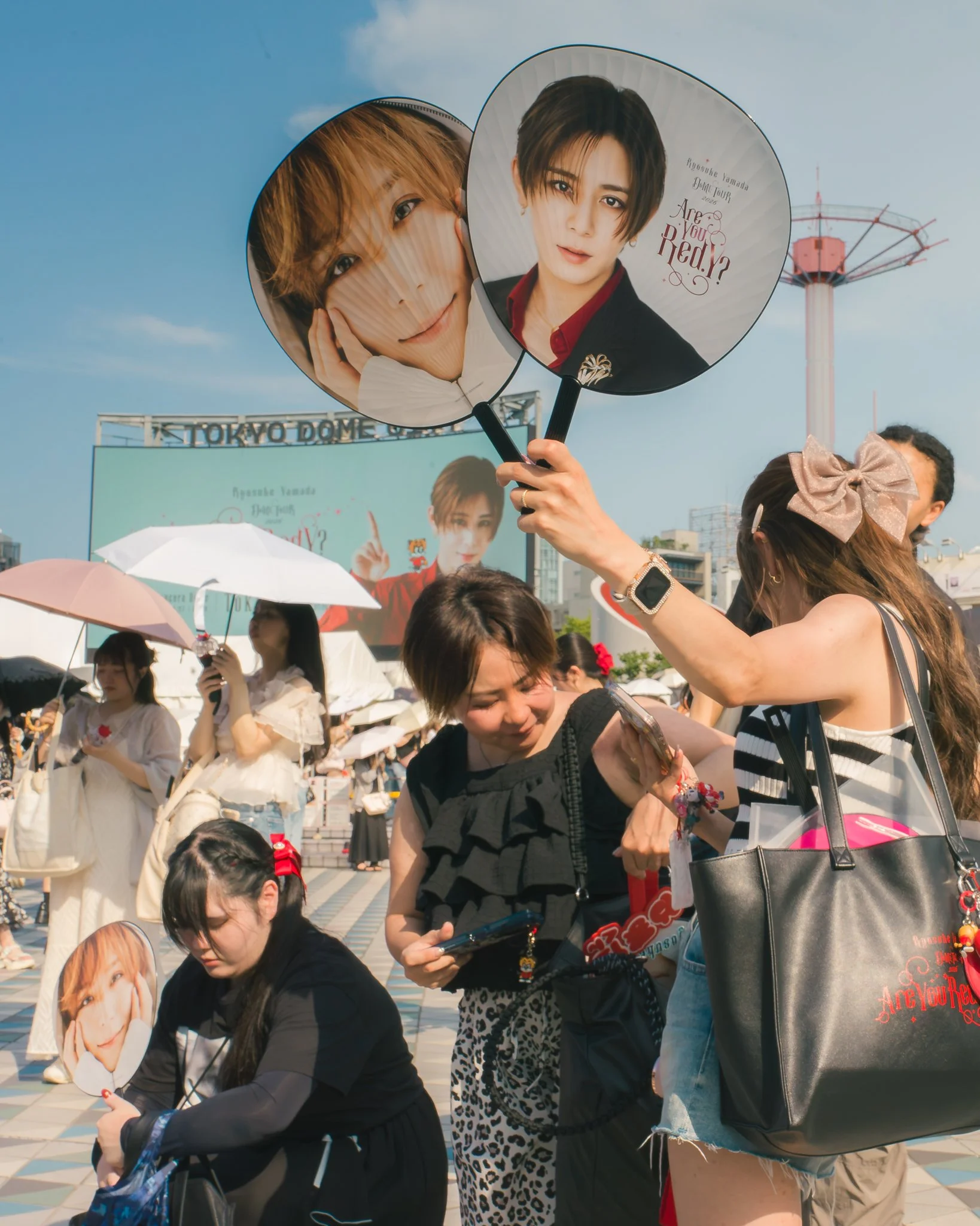 People holding fans featuring photos of a woman with short brown hair, and text reading 'Are You Red?' at an outdoor event near Tokyo Dome on a sunny day.