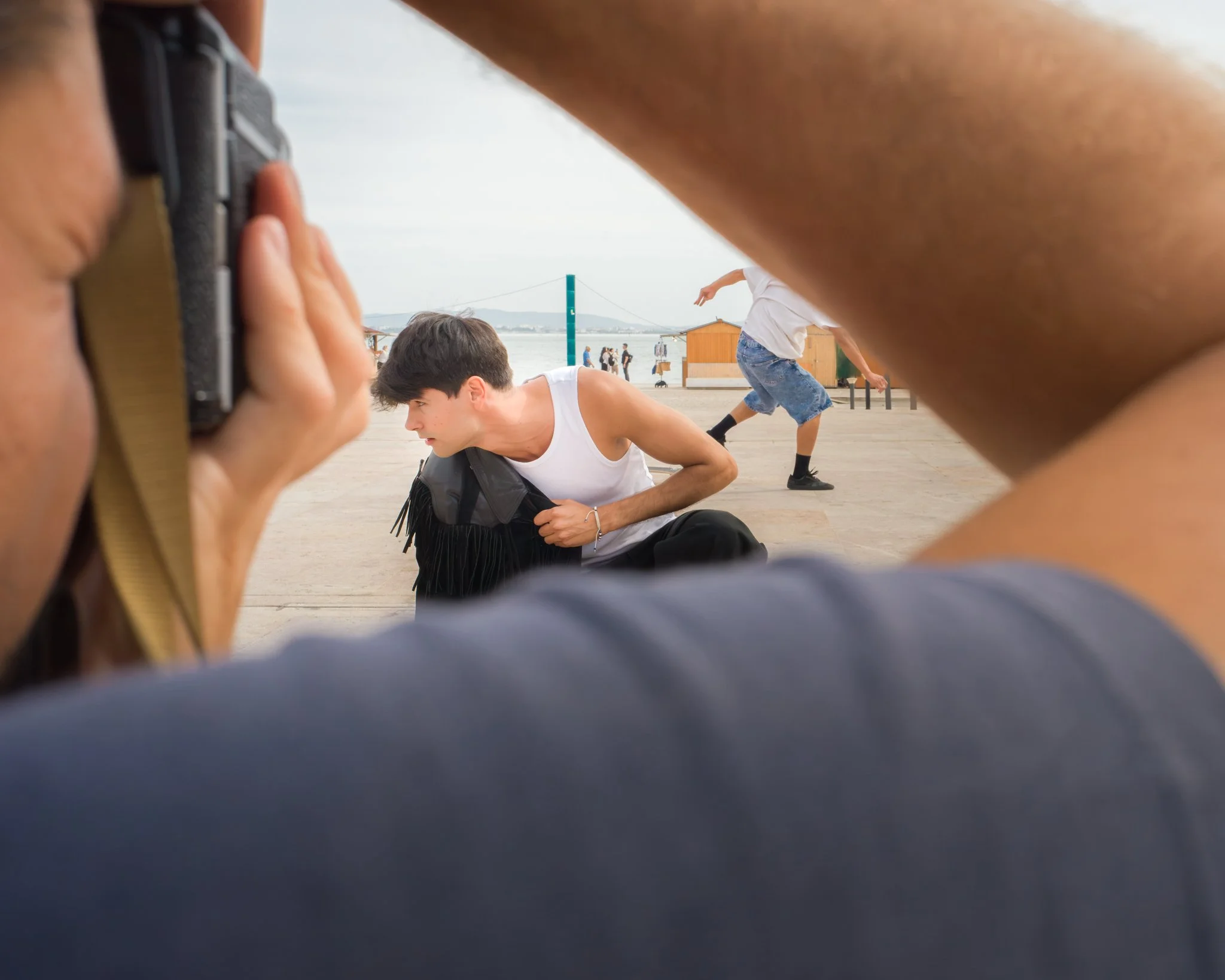 People taking a photo of a young man lying on the ground at a waterfront park, with other individuals and small buildings in the background.