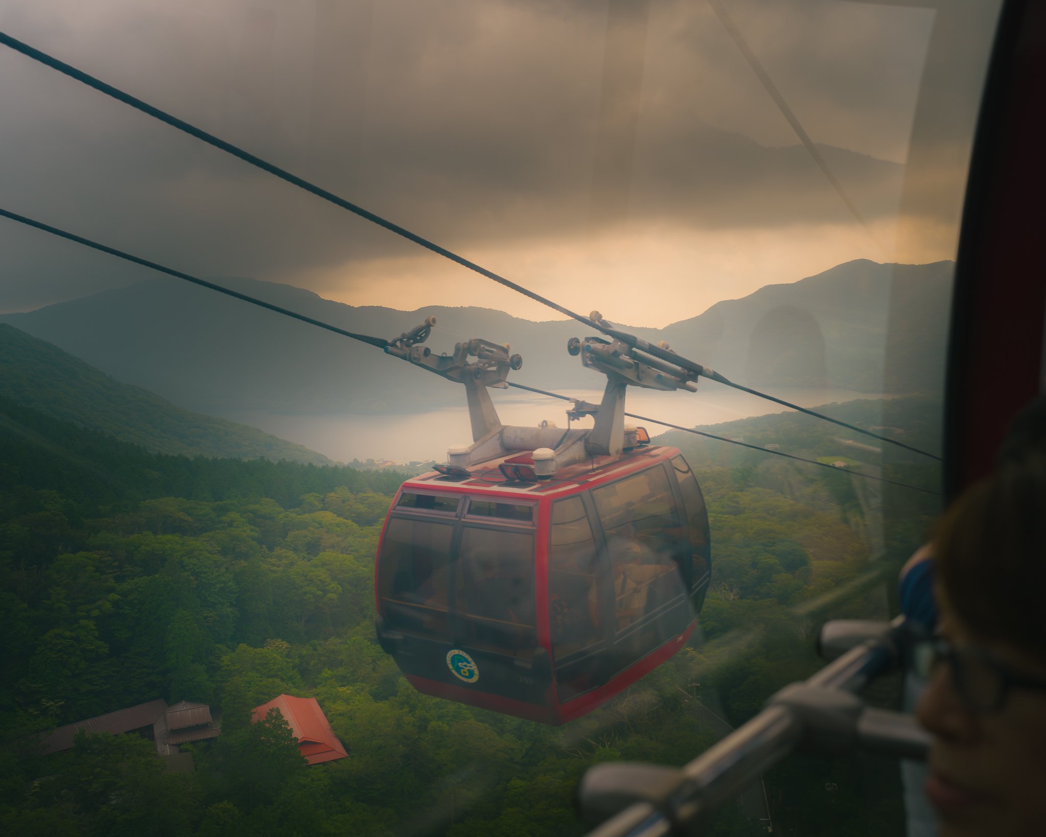 A panoramic view of a mountain landscape seen from inside a cable car with a red exterior, cable wires, and a cloudy sky overhead.