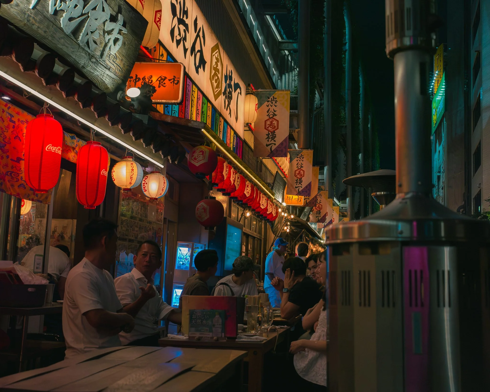 A busy night scene at an Asian street food outdoor seating area with red lanterns hanging and people sitting and eating, illuminated by colorful lights and signs with Asian characters.