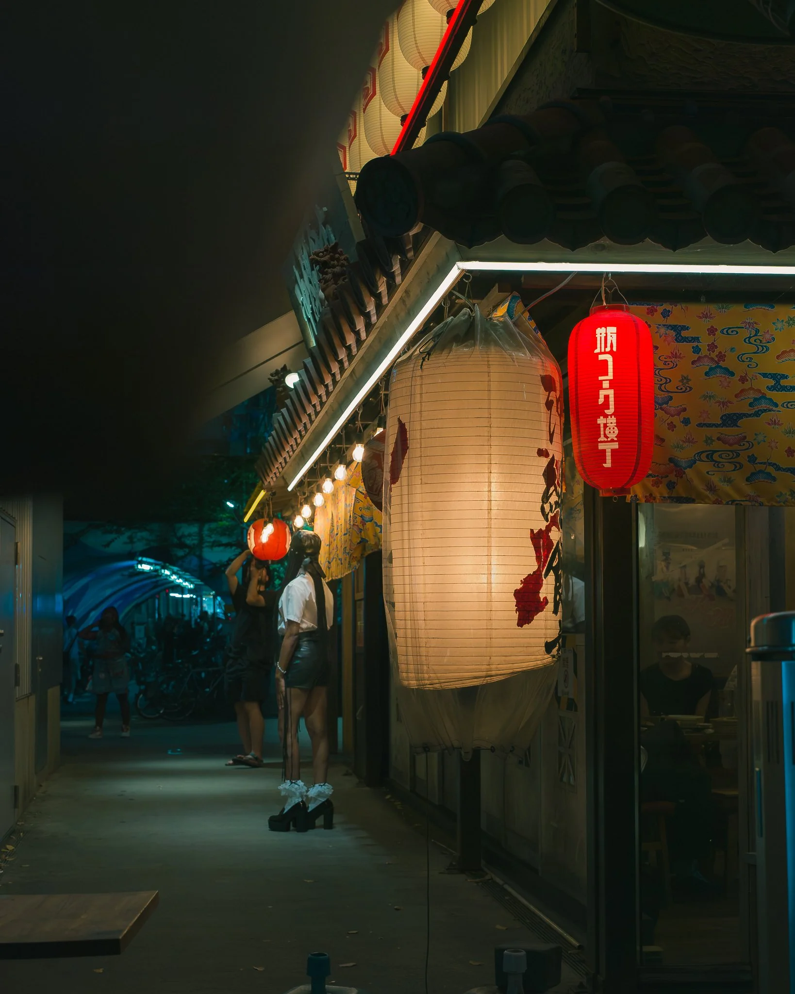 Night scene of a decorated street with lanterns, illuminated signs, and people walking, including a woman with a handbag standing near a large paper lantern with an Asian cityscape reflection on a glass surface.