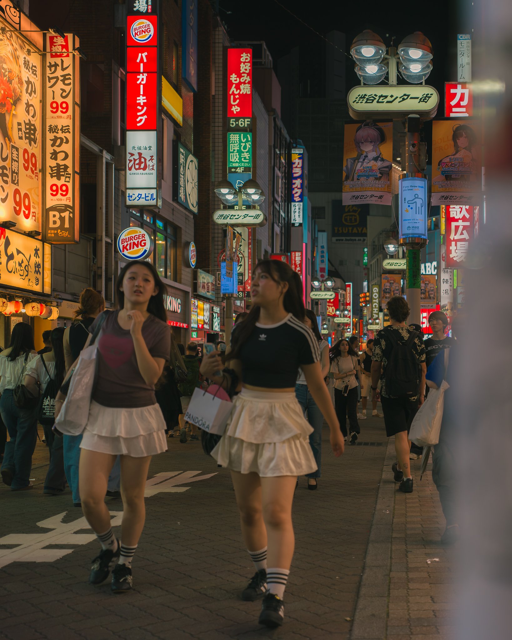 Nighttime crowded street in Japan with illuminated signs, pedestrians, two women in matching sporty outfits with skirts and striped socks, and various neon signs in Japanese.