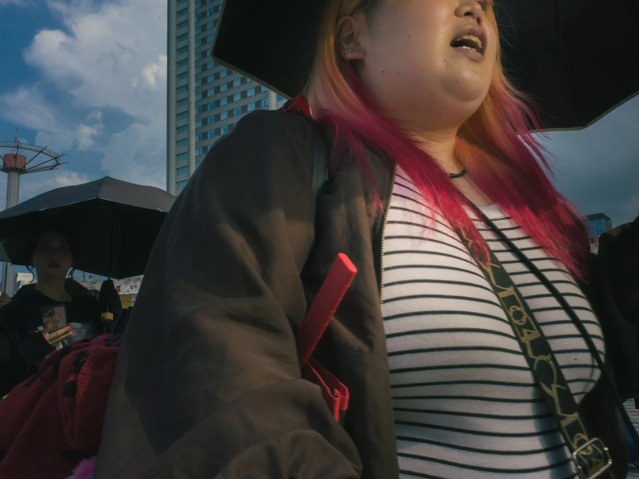 Close-up of a woman with pink hair and multiple ear piercings, wearing a striped shirt and black jacket, holding a pink object. In the background, there are tall buildings, a person holding an umbrella, and a partly cloudy sky.