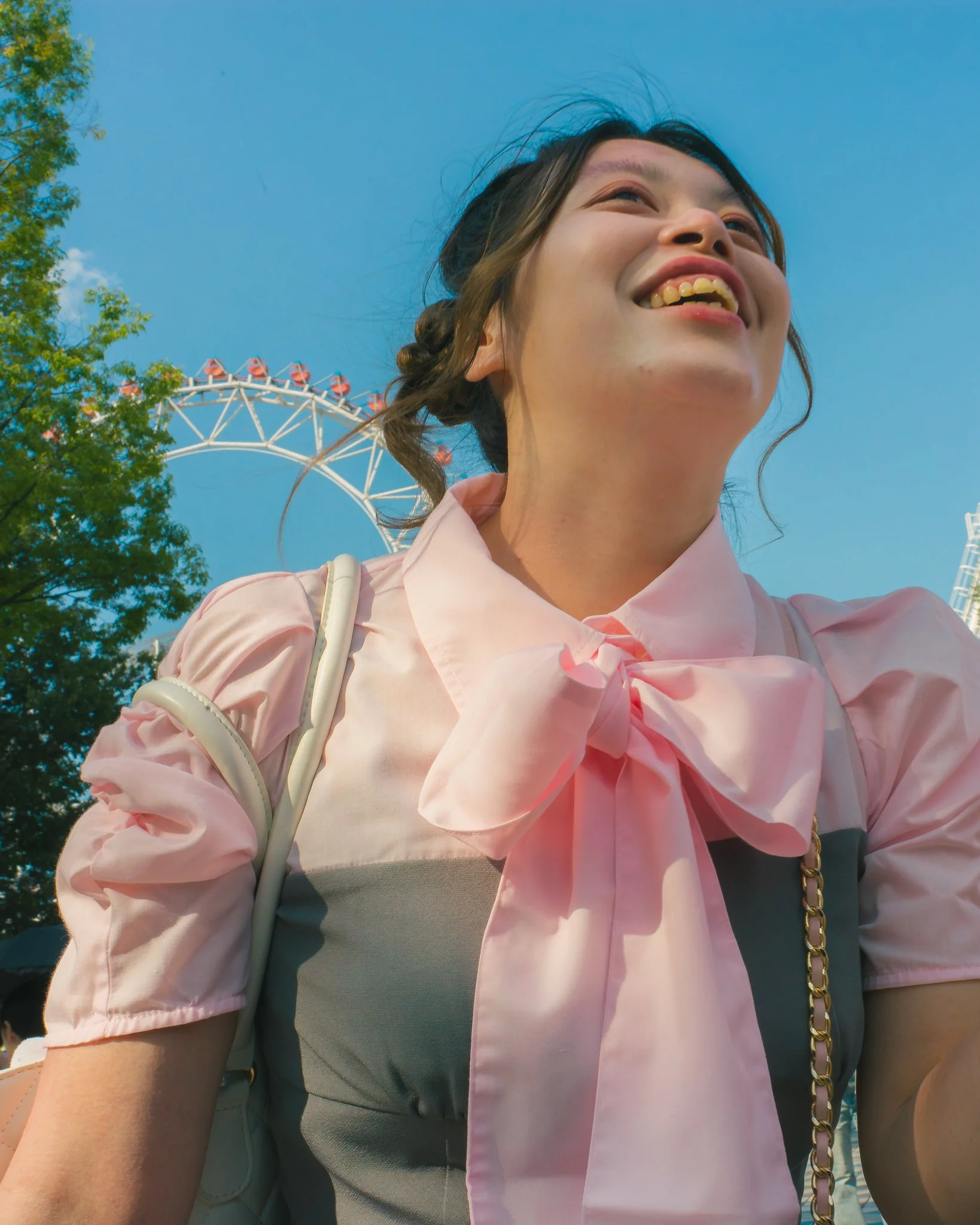 Close-up of a smiling woman with dark hair and a pink blouse, standing outdoors with a clear blue sky, trees, and a Ferris wheel in the background.