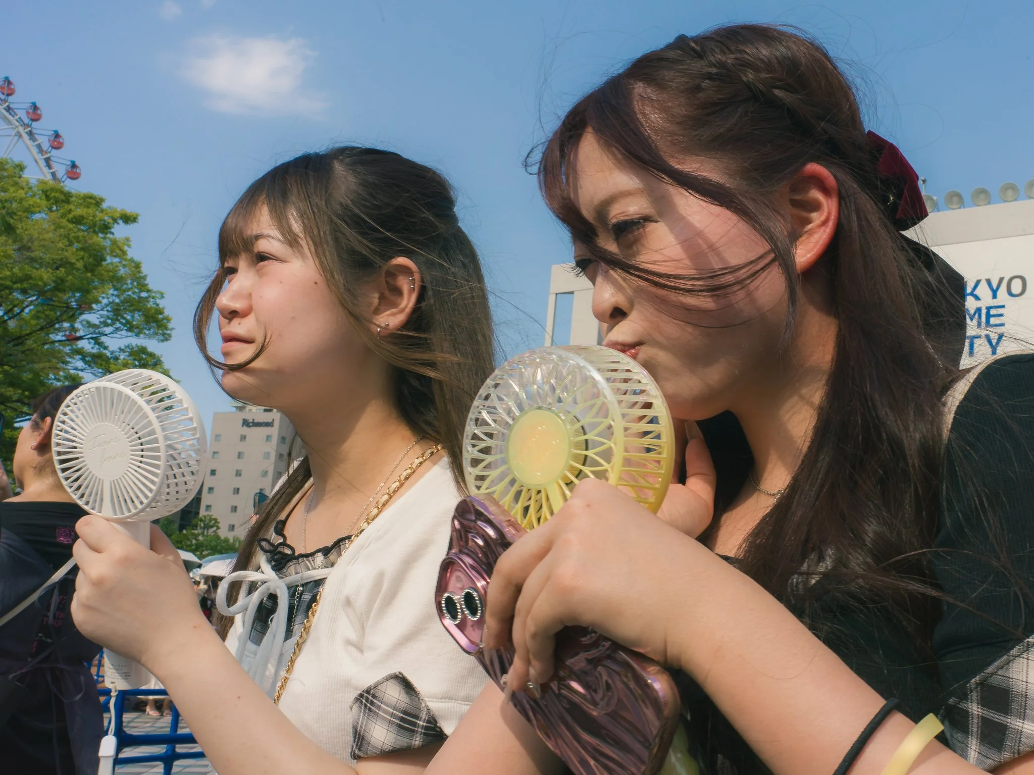 Two young women at an outdoor event, each holding a handheld fan and drinking from bottles, with a Ferris wheel and blue sky in the background.