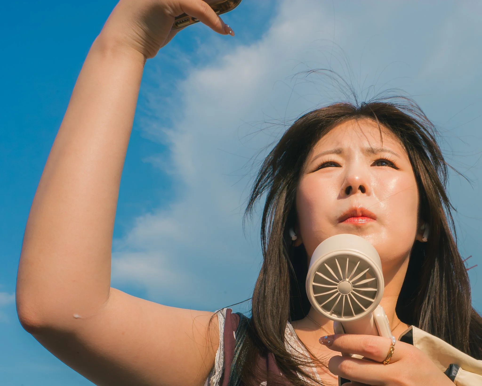 A young woman with long brown hair holding a vintage beige rotary telephone with a circular speaker at her chest, standing outdoors under a clear blue sky with a few clouds.