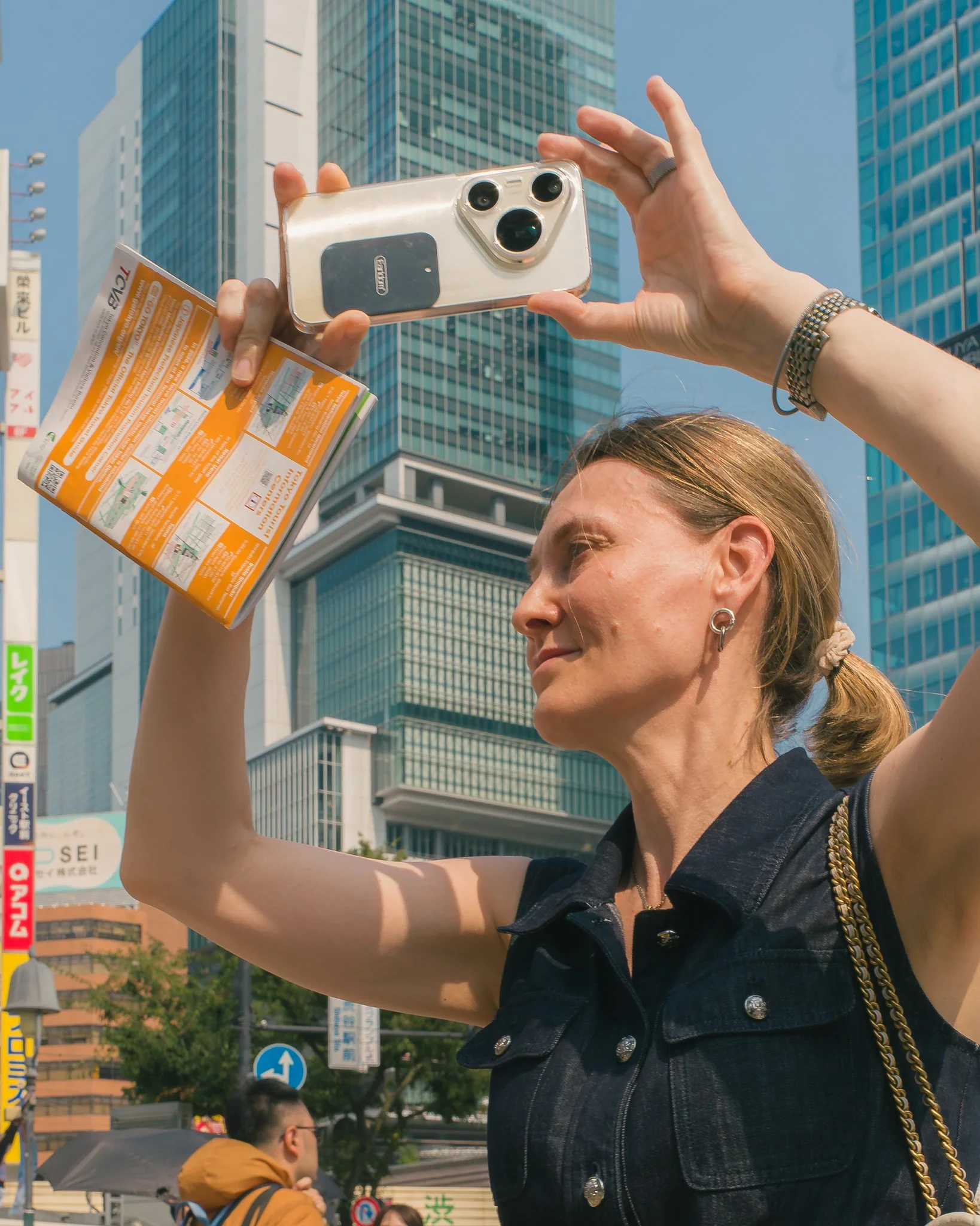 A woman is holding a smartphone with a transparent case and a ticket or coupon in an urban area. High-rise buildings and street signs are visible in the background. The woman has light brown hair tied back, and she is wearing a dark denim jacket with