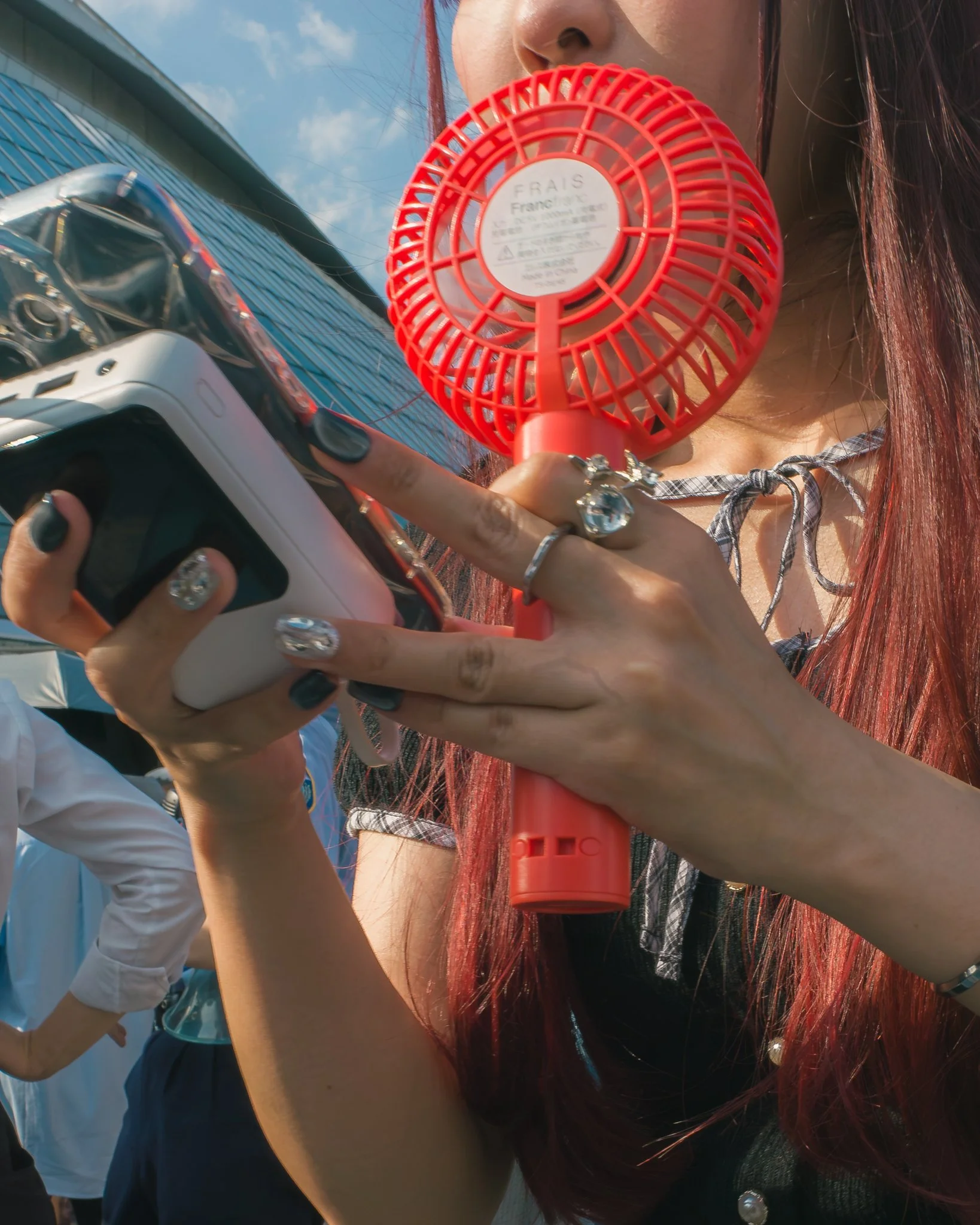 A young woman with red hair holding a small handheld fan close to her mouth, with a building and sky in the background.