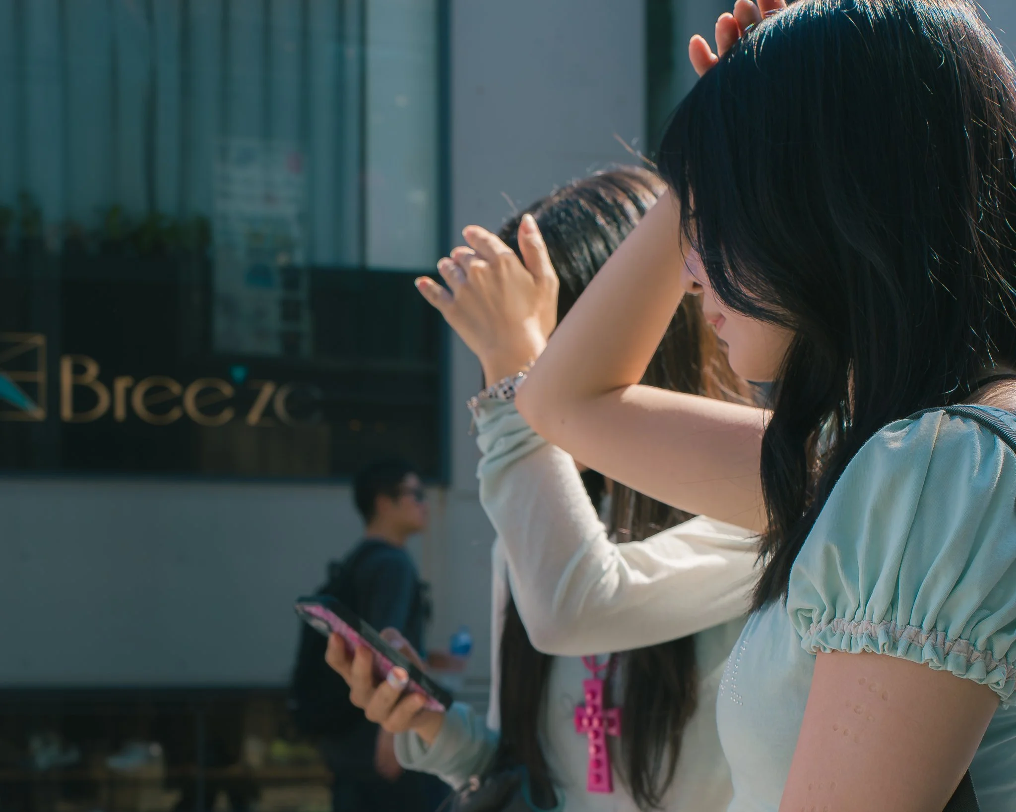 A person with dark hair is looking at their phone while standing outdoors. They are wearing a light-colored top with ruffled sleeves and a pink beaded necklace. Other people are visible in the background, some also using phones.
