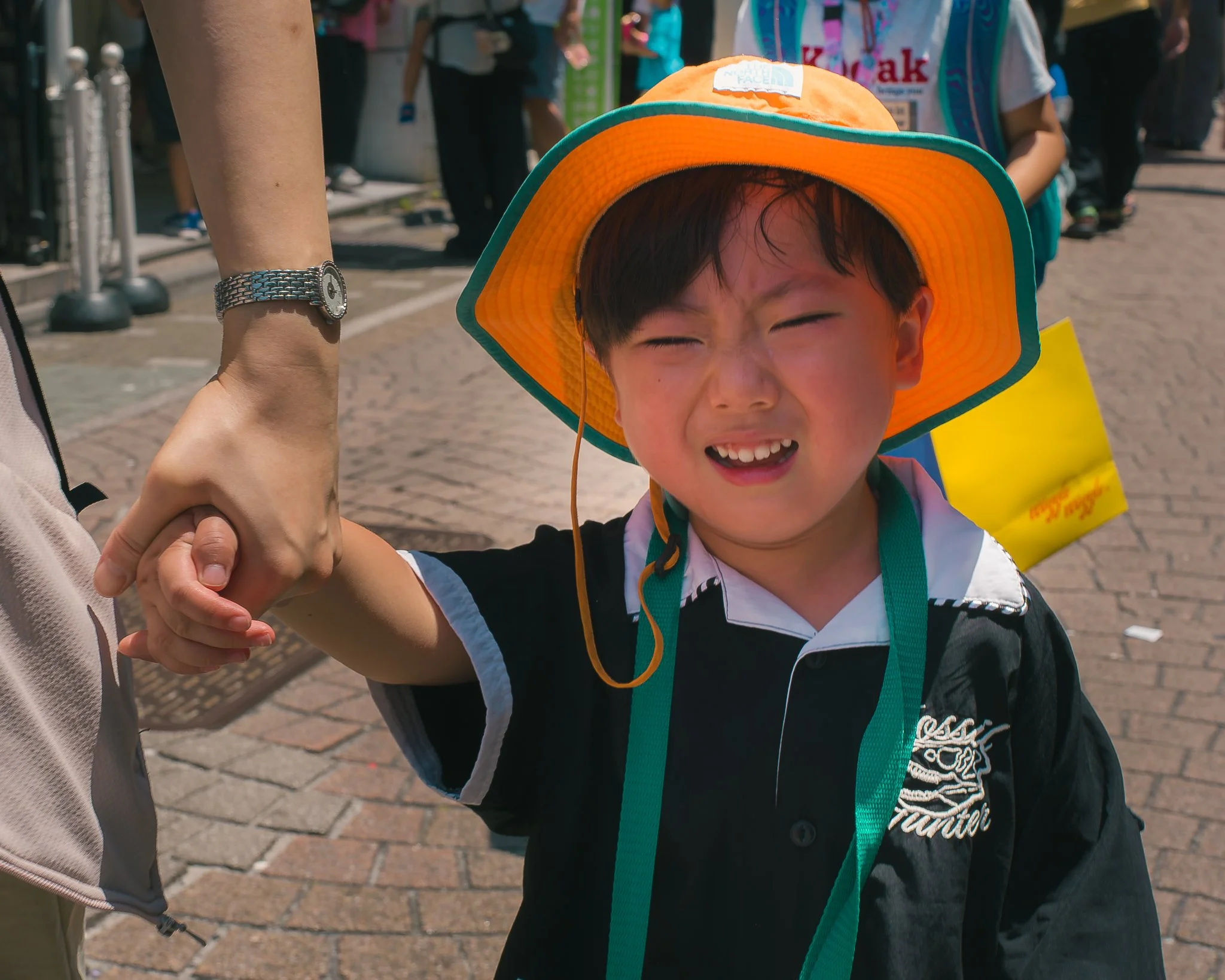 Close-up of a young boy with dark hair, wearing an orange hat with green trim, being held by an adult in a busy outdoor setting.