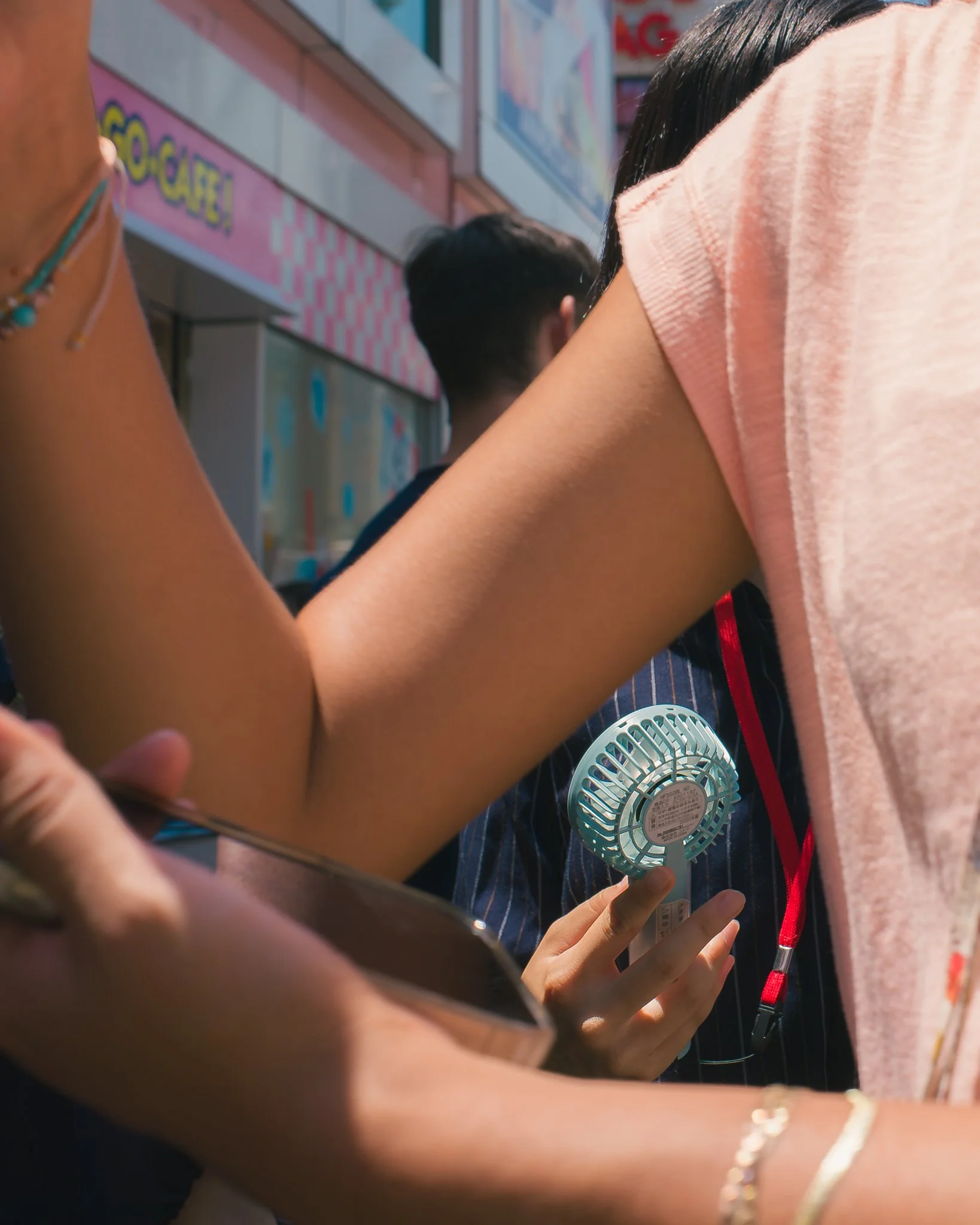 Close-up of a person holding a handheld fan in a crowded outdoor setting, with several other people around and colorful signage in the background.
