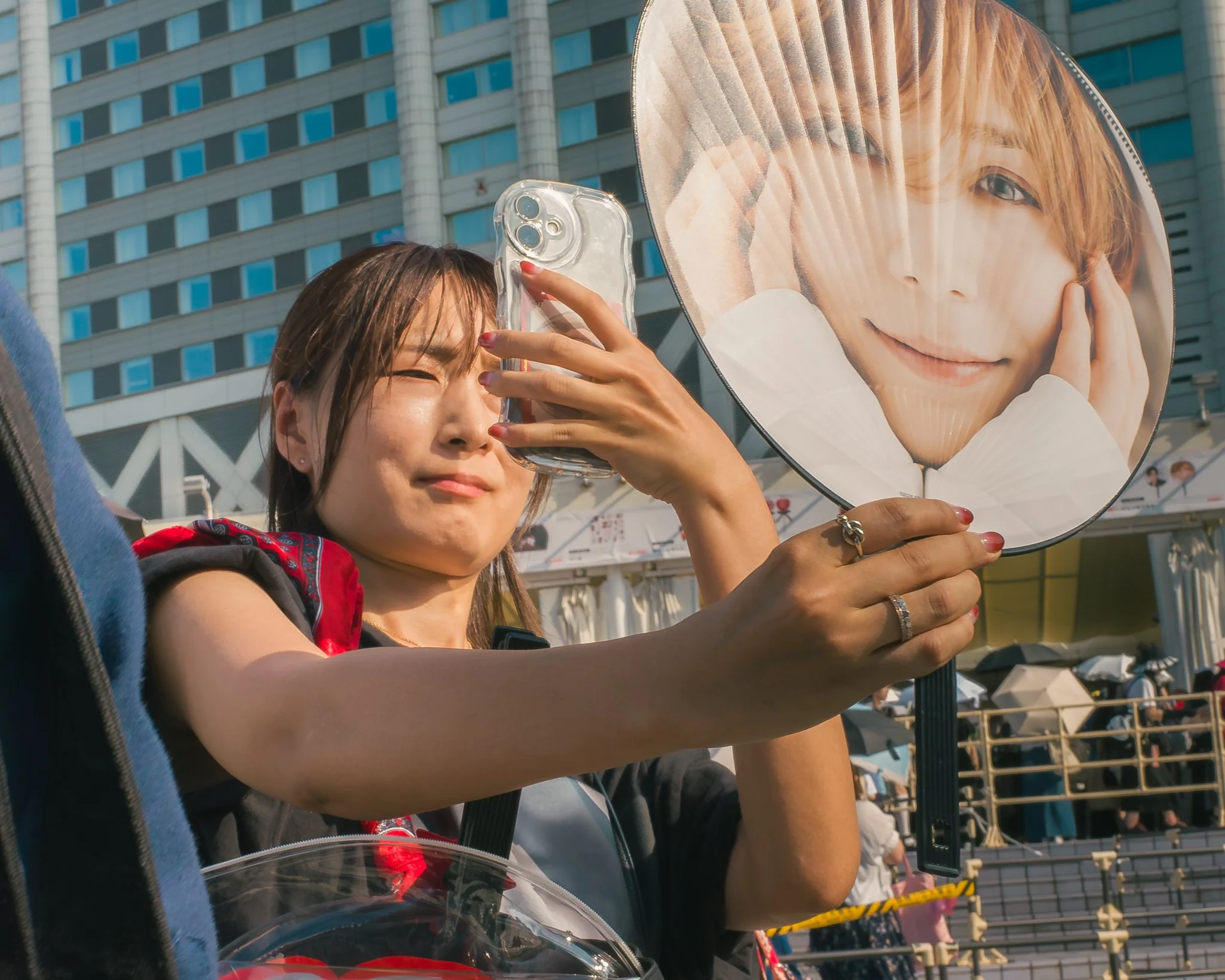 A woman is holding a large round fan with an image of a young person's face on it, while another woman is taking her photo with a smartphone. The scene is outdoors in front of a tall building with reflective windows.