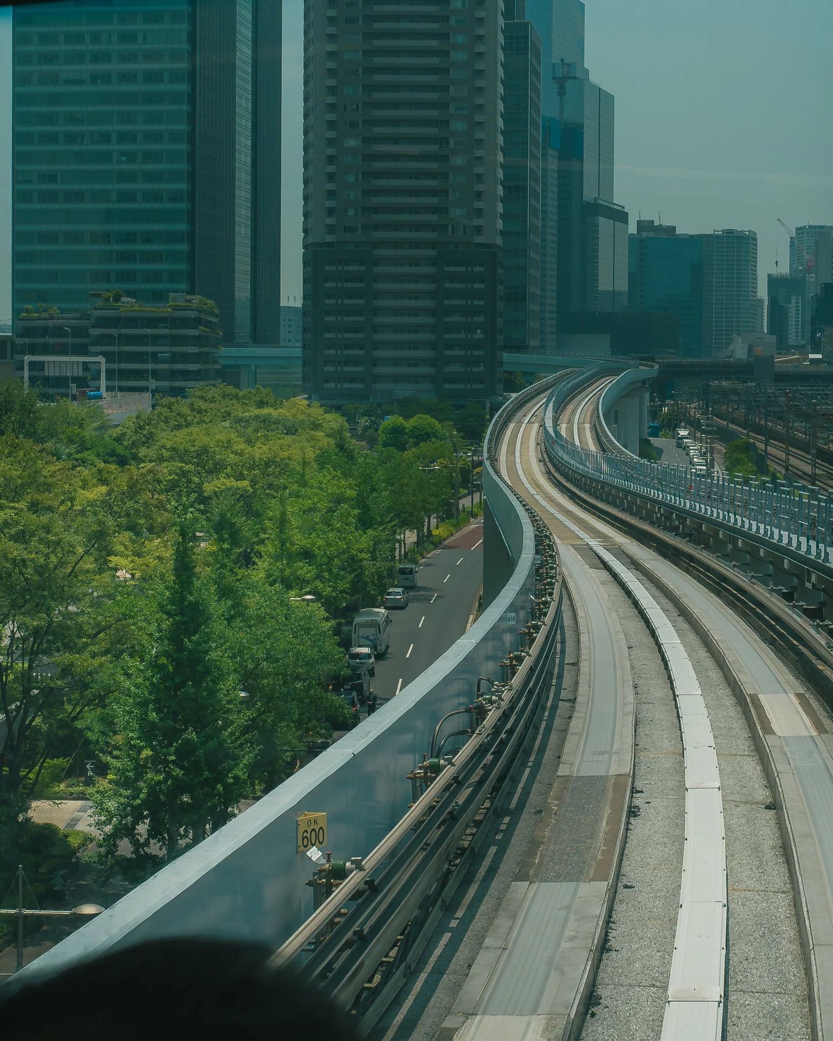 A cityscape with tall glass skyscrapers and a curved elevated train track in the foreground. There are green trees below the buildings and vehicles on the street. The sky appears hazy.