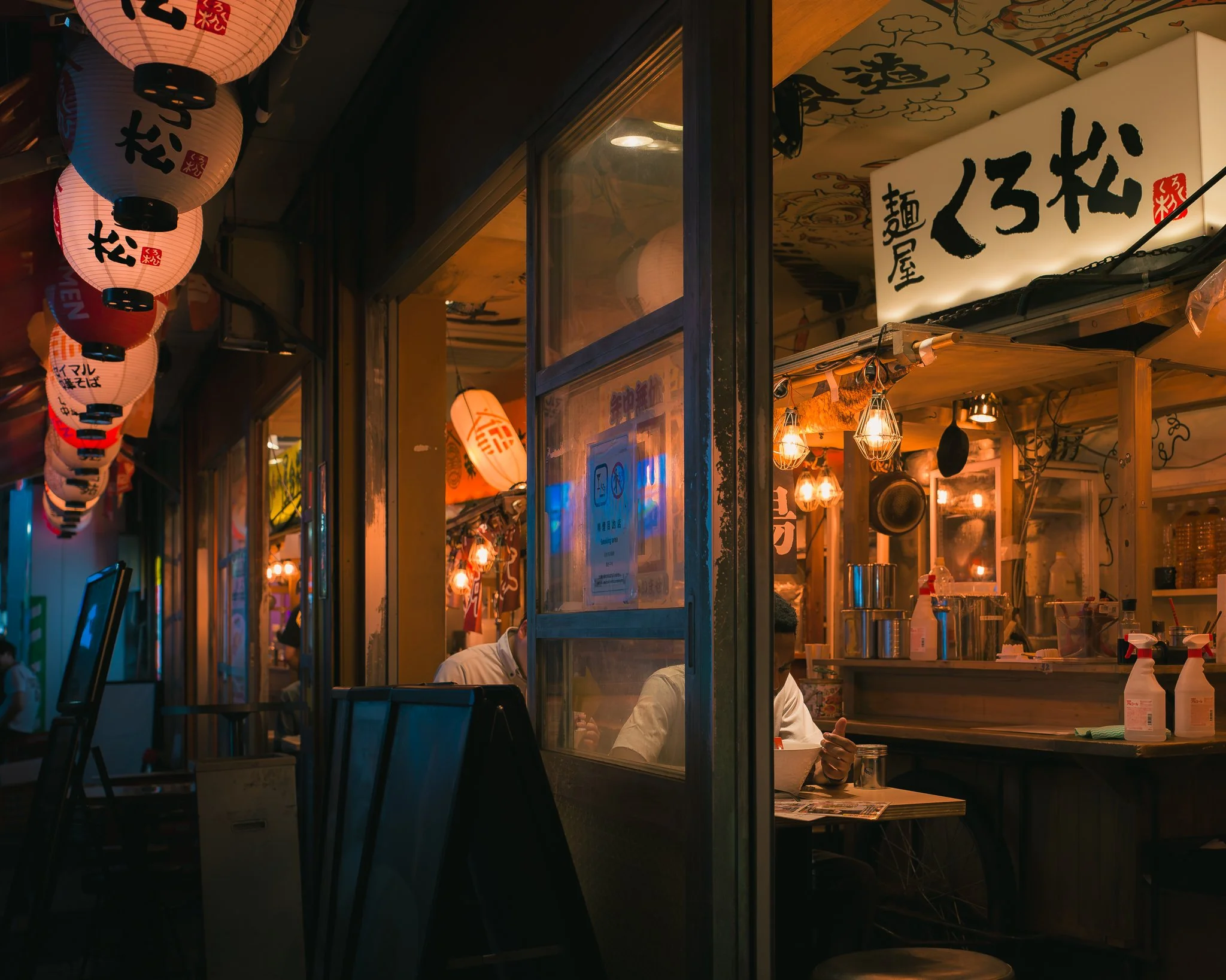 Night scene of a Japanese restaurant with traditional paper lanterns hanging outside, illuminated with warm light, and a window revealing a person inside preparing food.