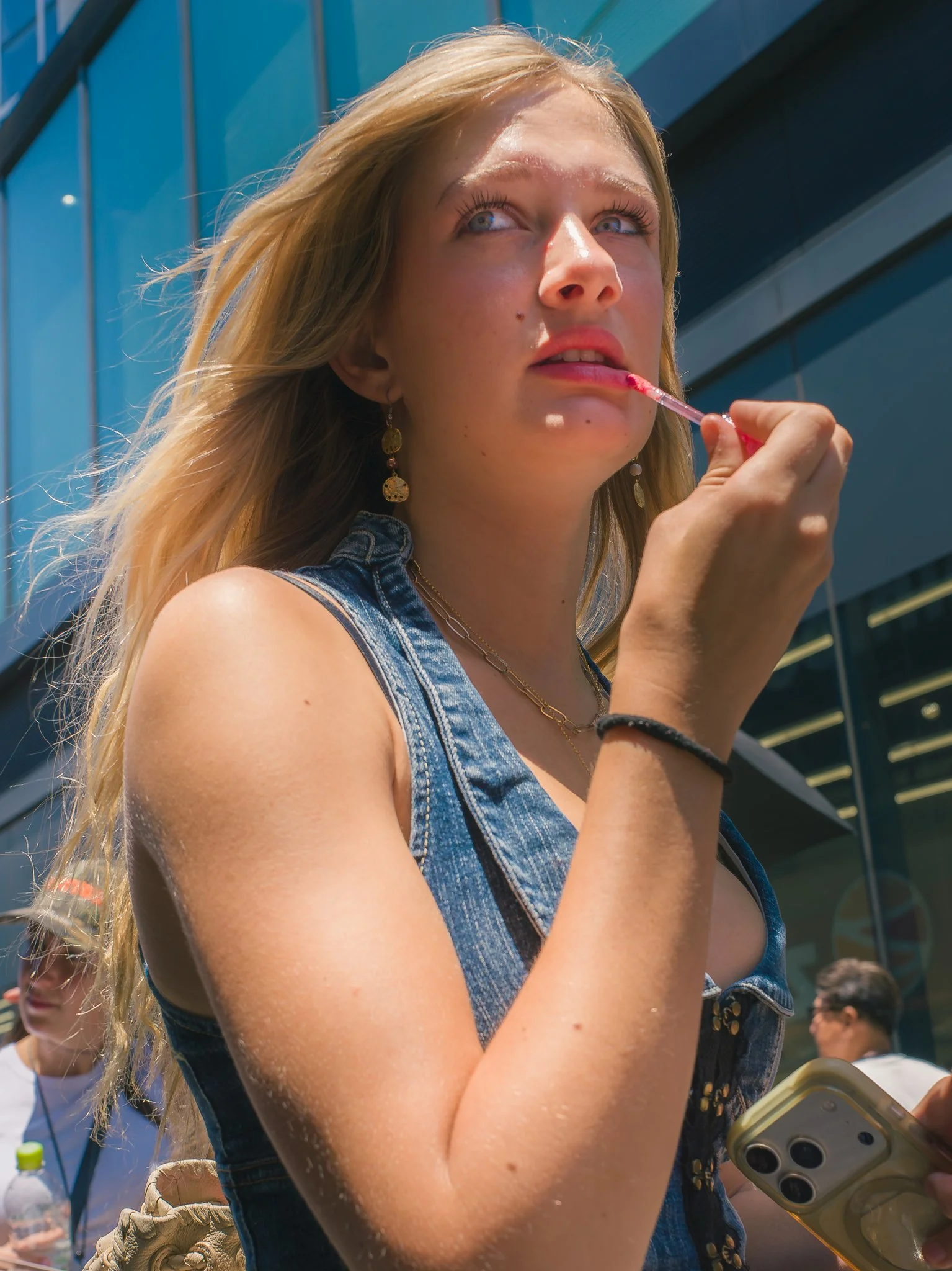 A young woman with blonde hair applying pink lipstick outside on a sunny day, holding a phone in her hand, in front of a modern building with reflective glass windows.