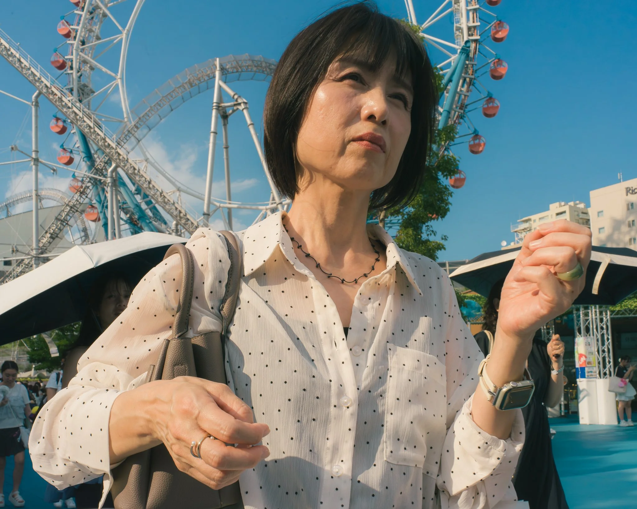 A woman wearing a white polka dot blouse with black necklace, holding her hands together, stands in front of a Ferris wheel during daytime. The sky is blue with few clouds, and other people are visible in the background, some holding umbrellas.